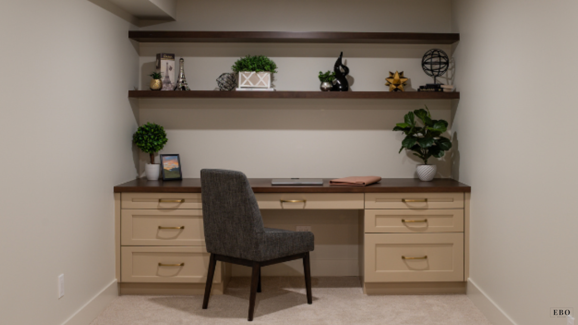 Built-in home office alcove featuring beige cabinets with gold handles, dark wood desktop, floating display shelves, and a gray upholstered chair.