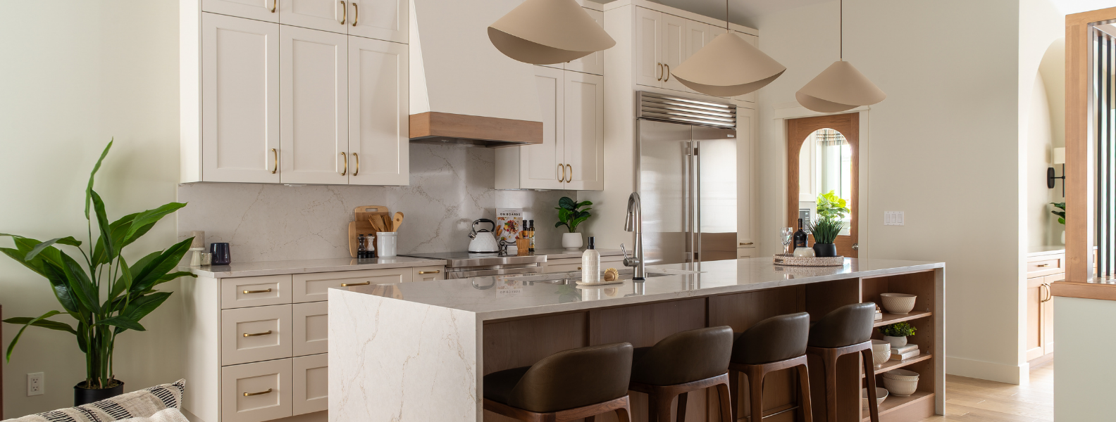 A modern kitchen with white cabinets, a large waterfall quartz island, and dark leather bar stools. Three unique pendant lights hang above. A sub-zero fridge and arched door are in the background.