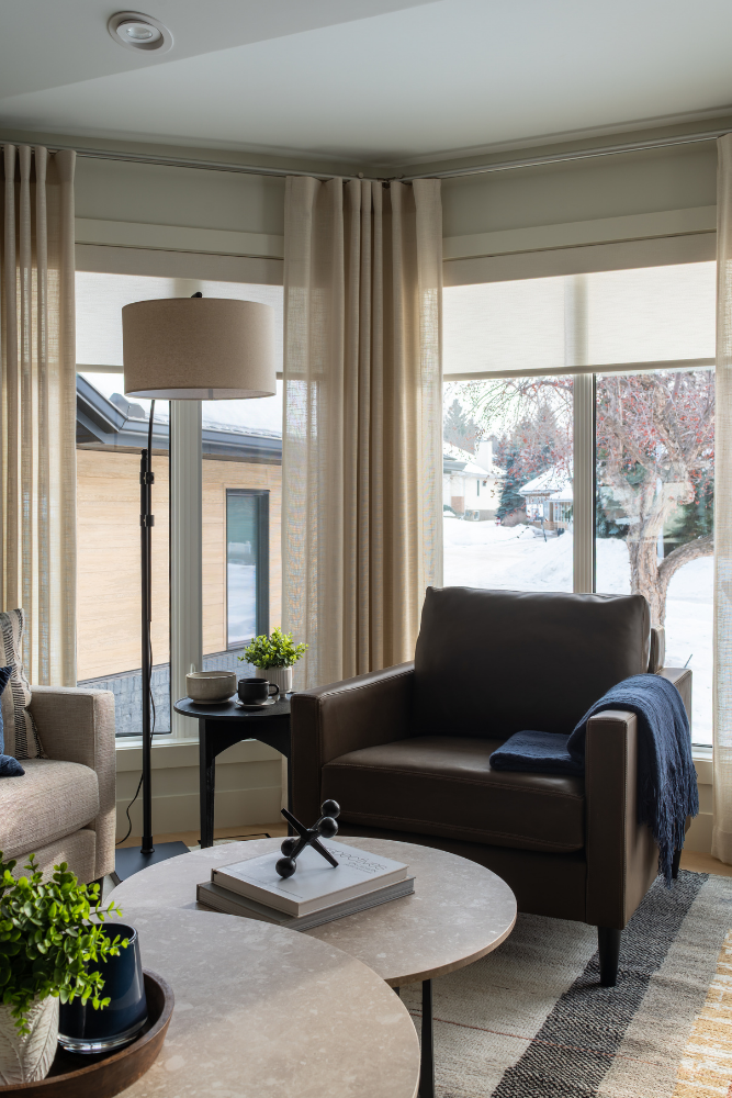 A focused shot of a cozy dark brown leather armchair with a navy blue throw blanket, positioned next to a modern black arc floor lamp in a light-filled living room.