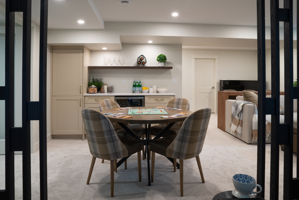 A wide-angle view of a well-appointed basement space, framed by dark mullion partitions in the foreground. It features a round game table surrounded by plaid chairs, a full wet bar with shelving, and a couch.