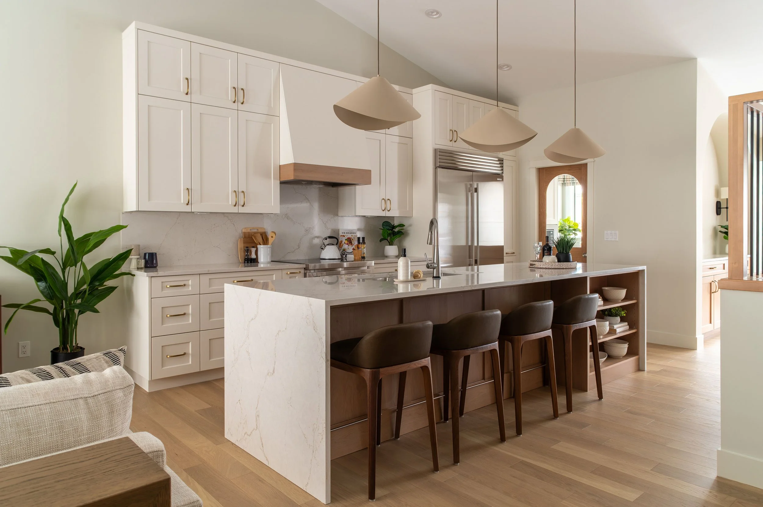 Warm neutral kitchen renovation by an Edmonton interior designer featuring white shaker cabinetry, brass hardware, quartz waterfall island, wood open shelving, and modern pendant lighting in a bright, family-focused open-concept home.