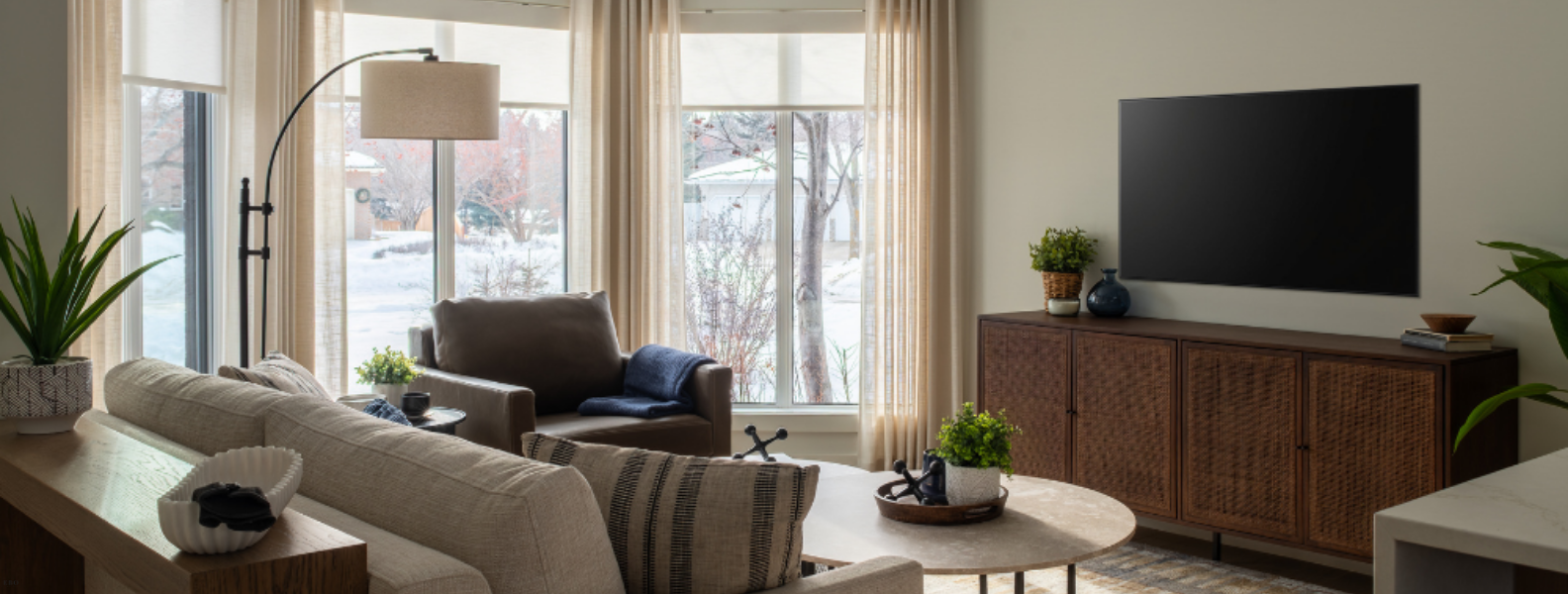 Cozy modern living room featuring a neutral sectional sofa, brown cane media console under a wall-mounted TV, and floor-to-ceiling windows with sheer drapes.
