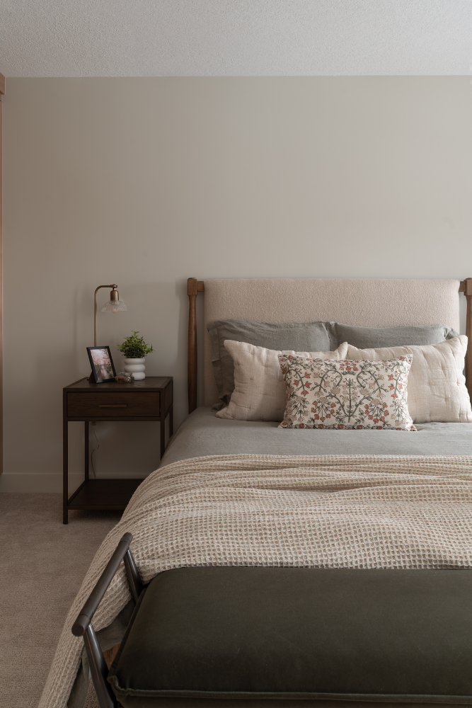 A close-up view of a cozy bed with an upholstered headboard, dressed in layers of textured linen bedding in cream, beige, and sage green, including a floral decorative pillow. A wood nightstand and lamp sit to the left.