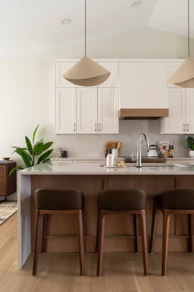 A modern kitchen featuring white shaker cabinets, a waterfall-edge marble island with three leather barstools, and conical beige pendant lights.