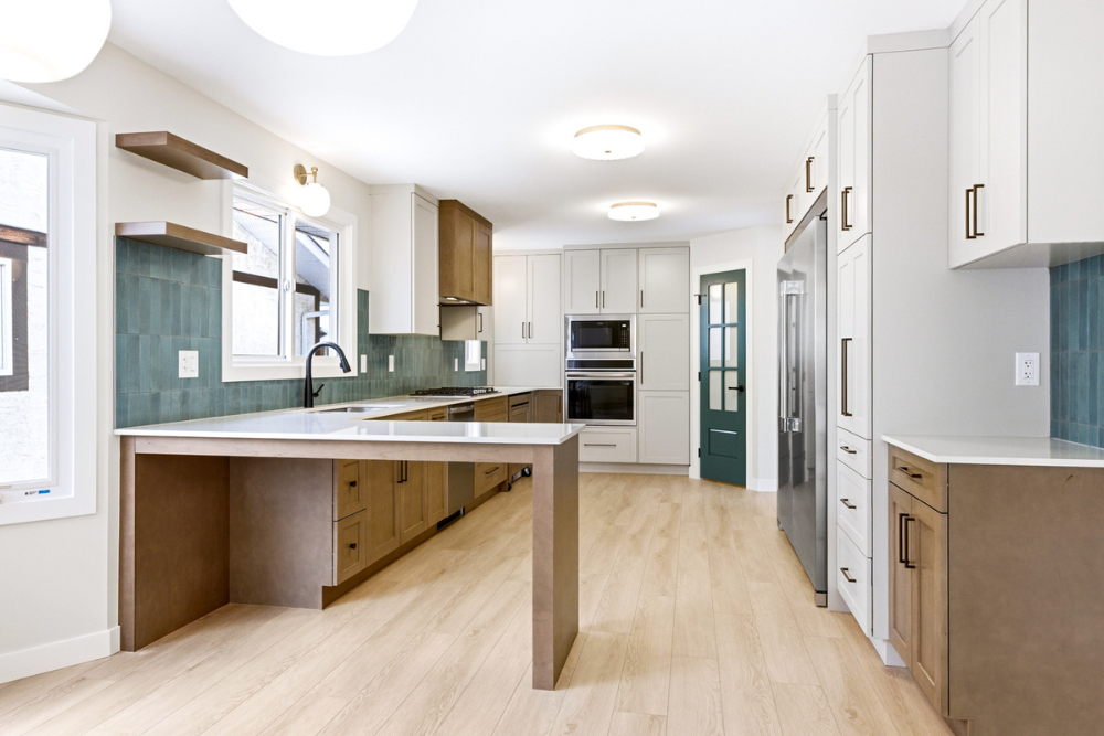 A spacious open-concept kitchen with light grey upper cabinets, medium-toned wood lower cabinets, white countertops, and a peninsula. A dark teal door with glass panes is visible in the background.