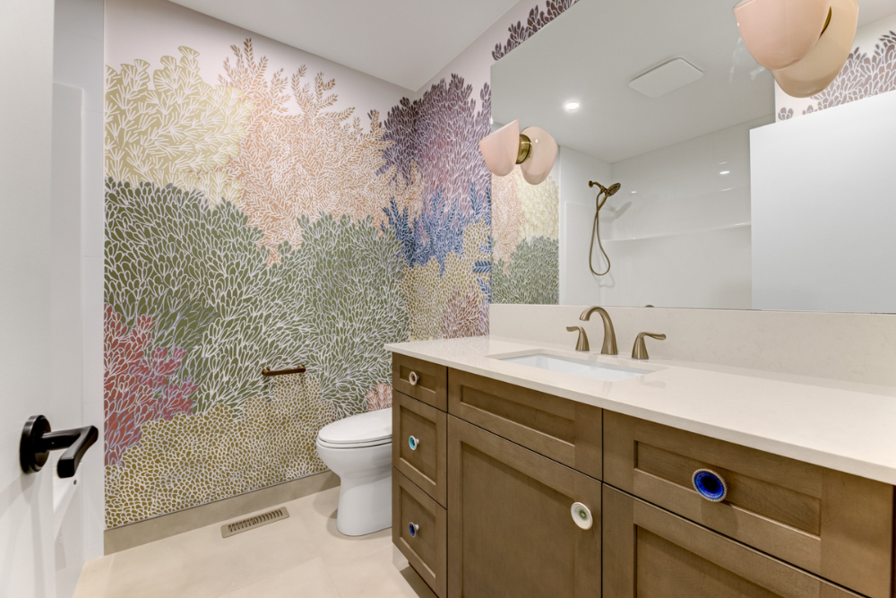 A unique powder room with a full-wall botanical pattern wallpaper in muted pastel colors (corals, greens, purples). It features a custom light wood vanity with decorative round knobs, two globe-shaped pink wall sconces above a large mirror, a white t