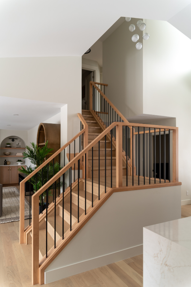 A photograph taken from above a kitchen island, looking down a natural wood staircase with a sleek black and oak railing in a modern, open-plan home.