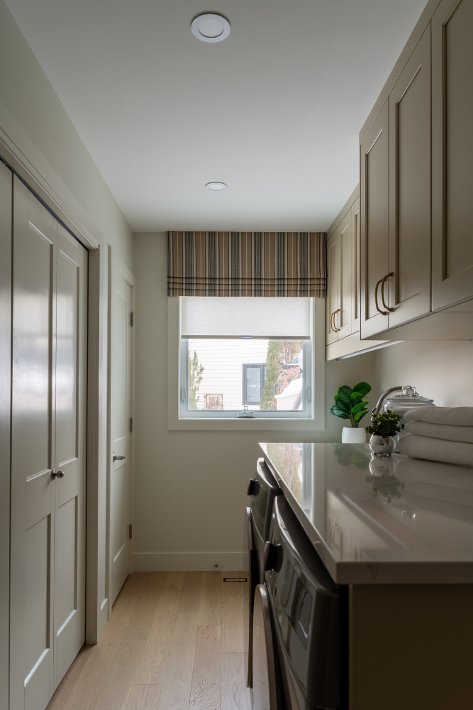 A narrow laundry room with light beige cabinetry, white front-load appliances, a quartz counter, and a window with a patterned roman shade overlooking a winter street.