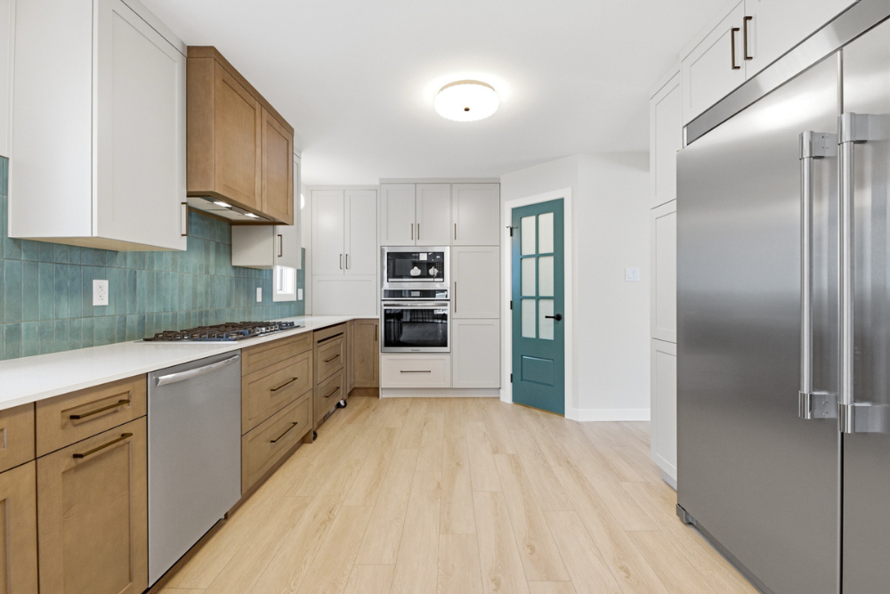A modern kitchen with medium-toned wood lower cabinets, light grey upper cabinets, a teal square-tile backsplash, and a stainless steel gas cooktop. Next to the cooking area are double stainless steel wall ovens and a dark teal painted door with glas
