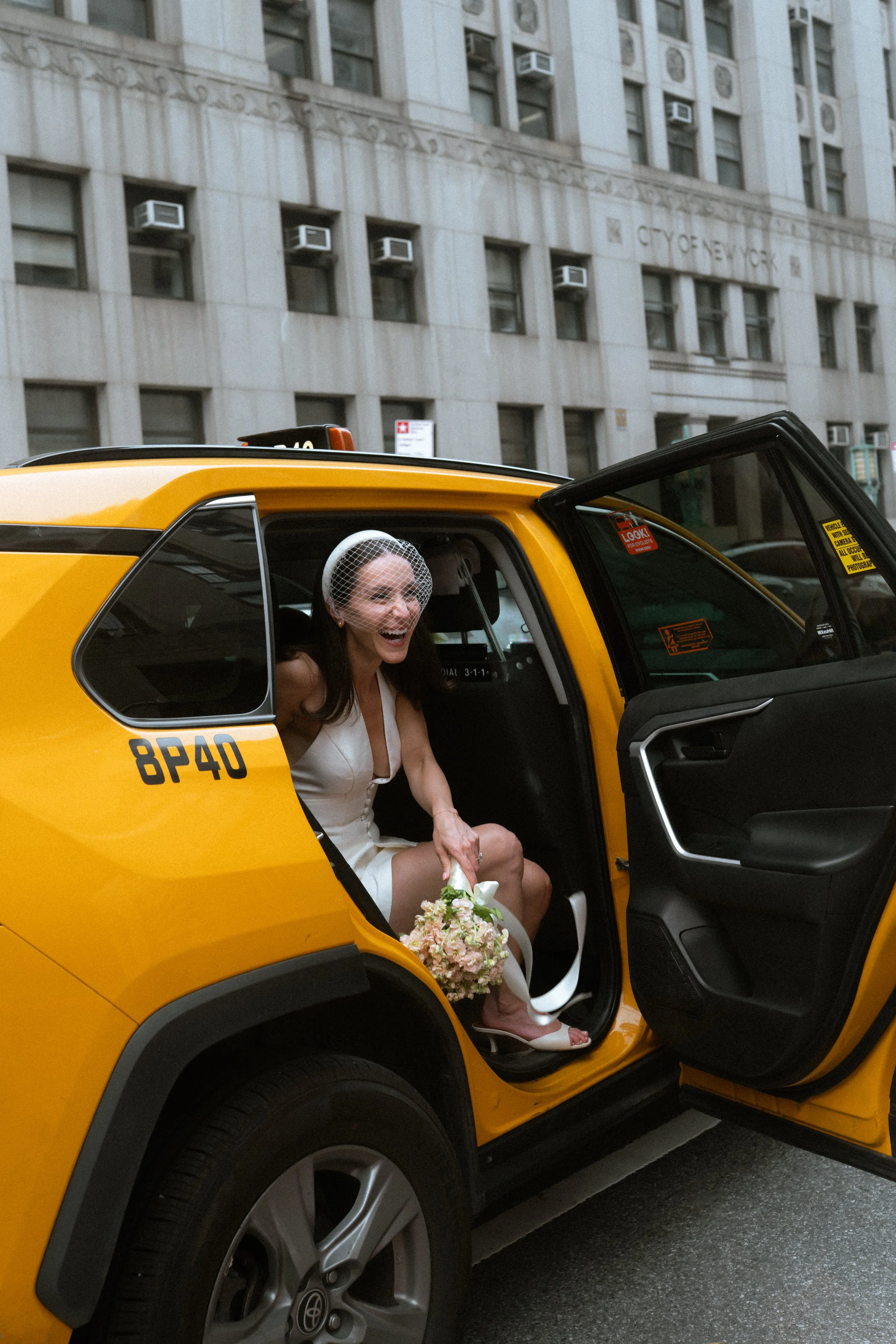 A woman in a white dress with a birdcage veil sitting inside a yellow taxi cab, holding a bouquet of pink and white flowers, smiling nyc soho city clerk manhattan elopement by jordan niccola