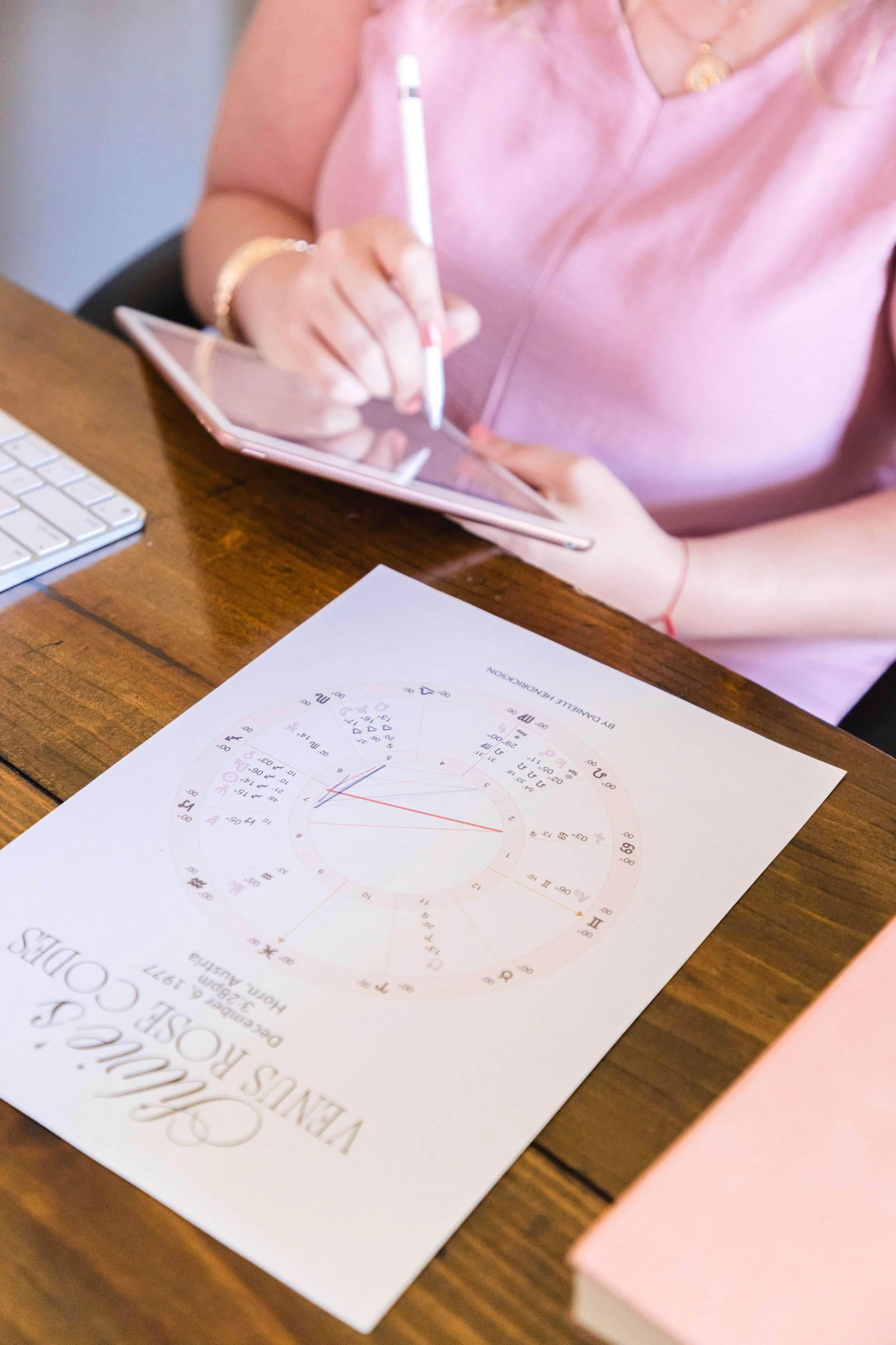 A woman in a pink shirt is using a stylus on a tablet at a wooden desk, with a printed astrological chart titled 'Venus in the Houses & Decan' and a white keyboard nearby.