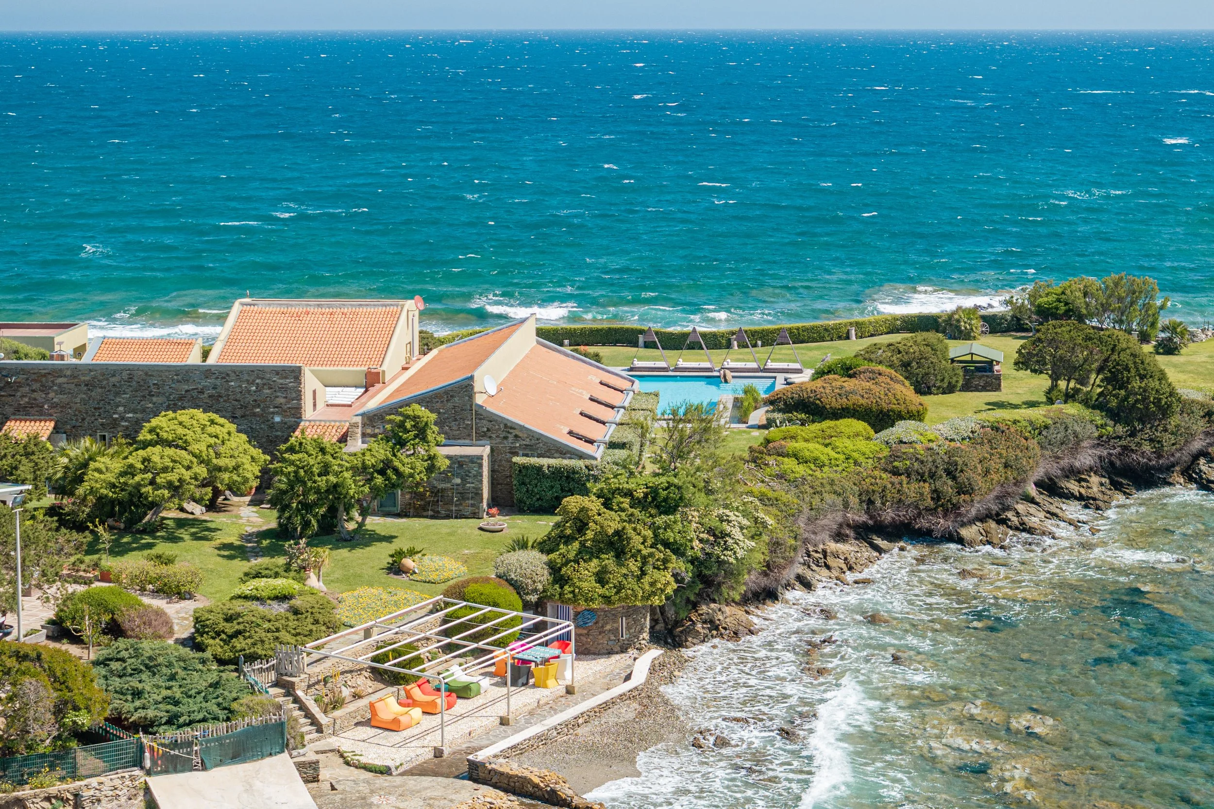 Aerial view of a seaside property with a swimming pool, lush greenery, and a rocky shoreline leading to the ocean.