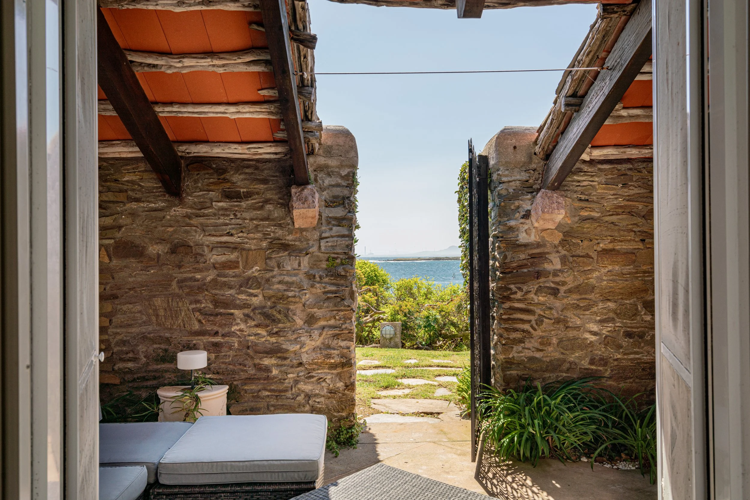 View through an open doorway showing a stone wall patio with a cushioned outdoor lounge, a small potted plant with a lamp on top, green plants, and a view of water and sky in the background.