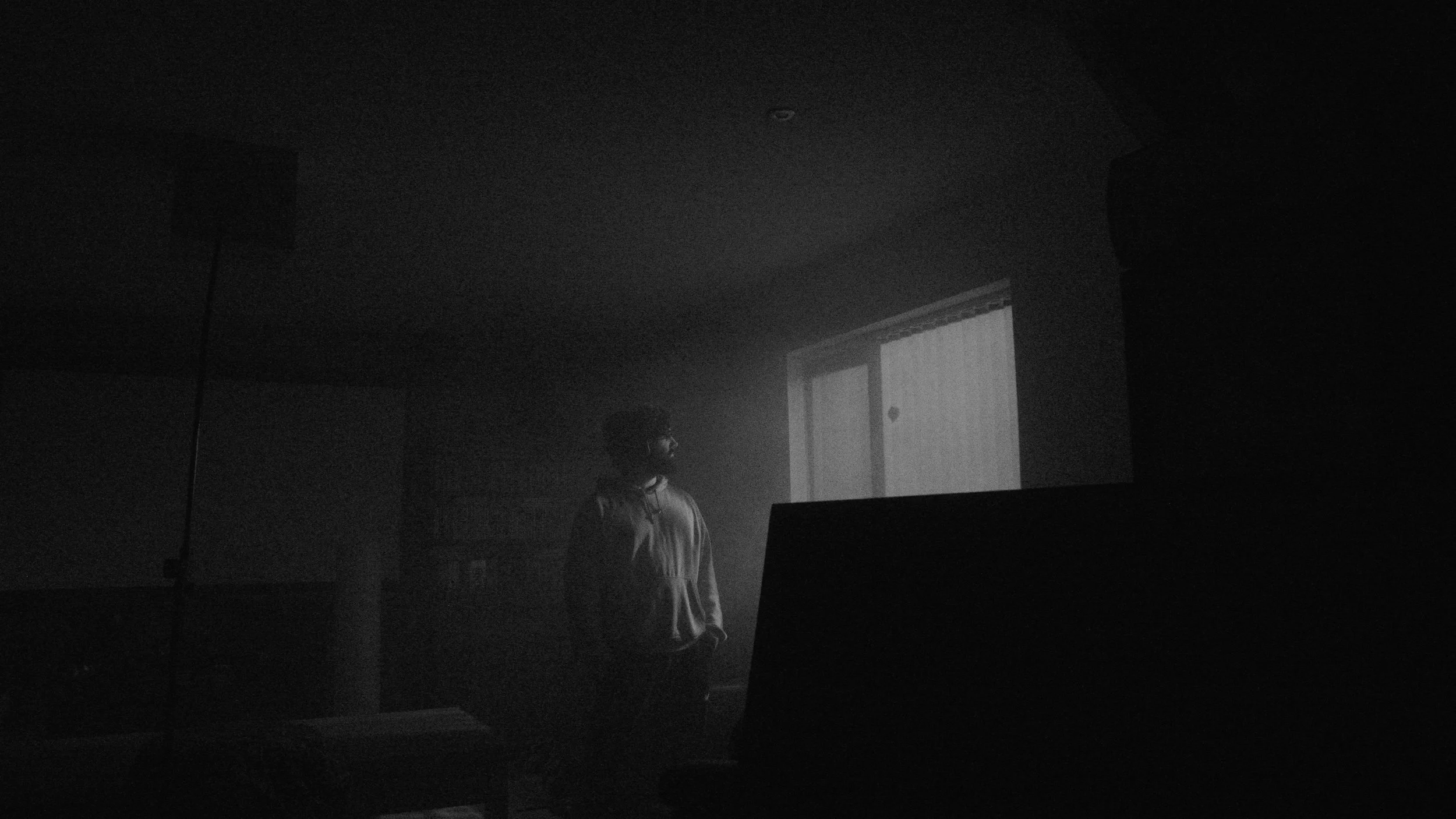 A person standing in a dark room with a faint light illuminating a screen or window behind them.
