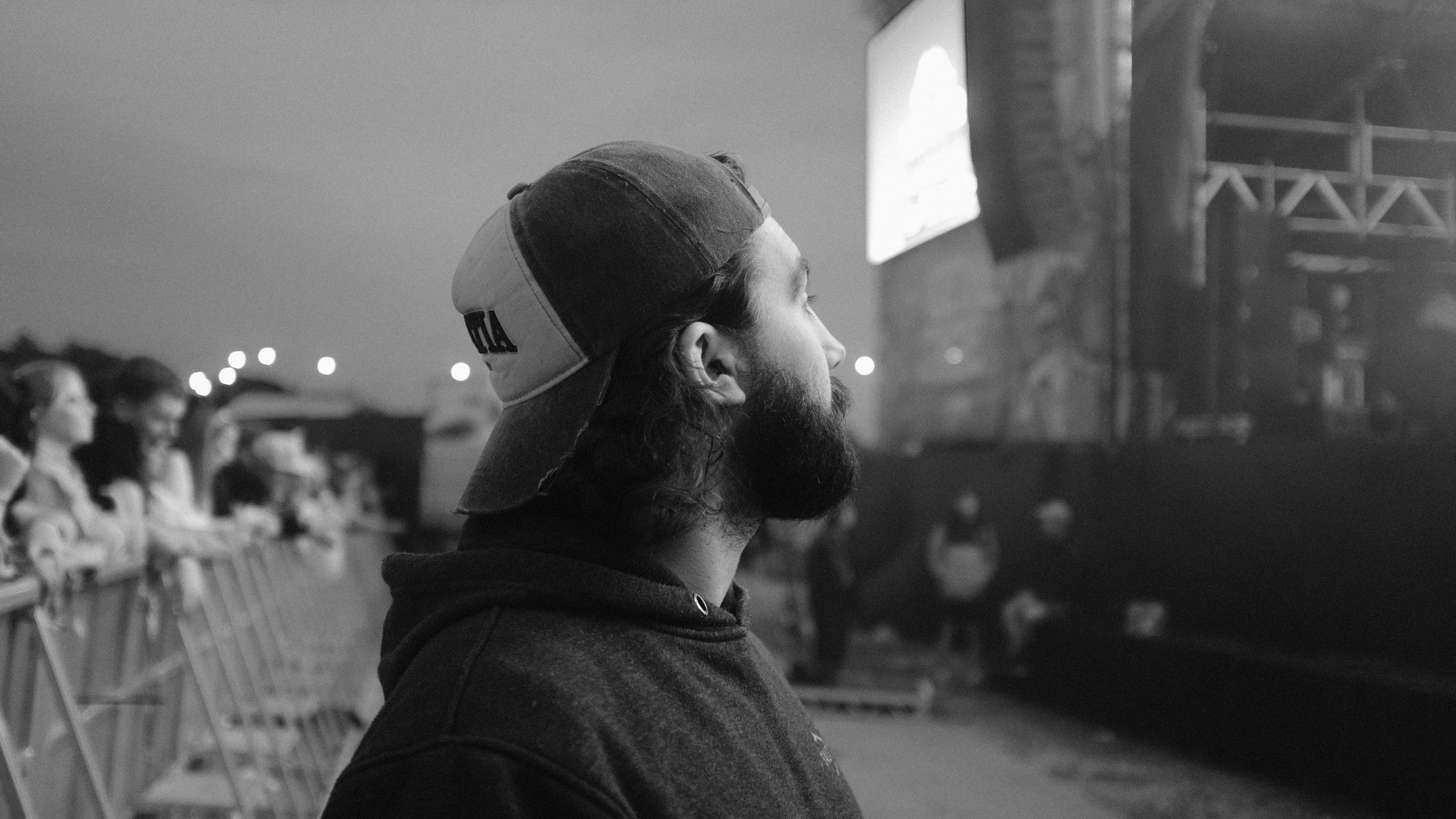 A man with a beard and long hair wearing a cap and hoodie, watching an event at night.