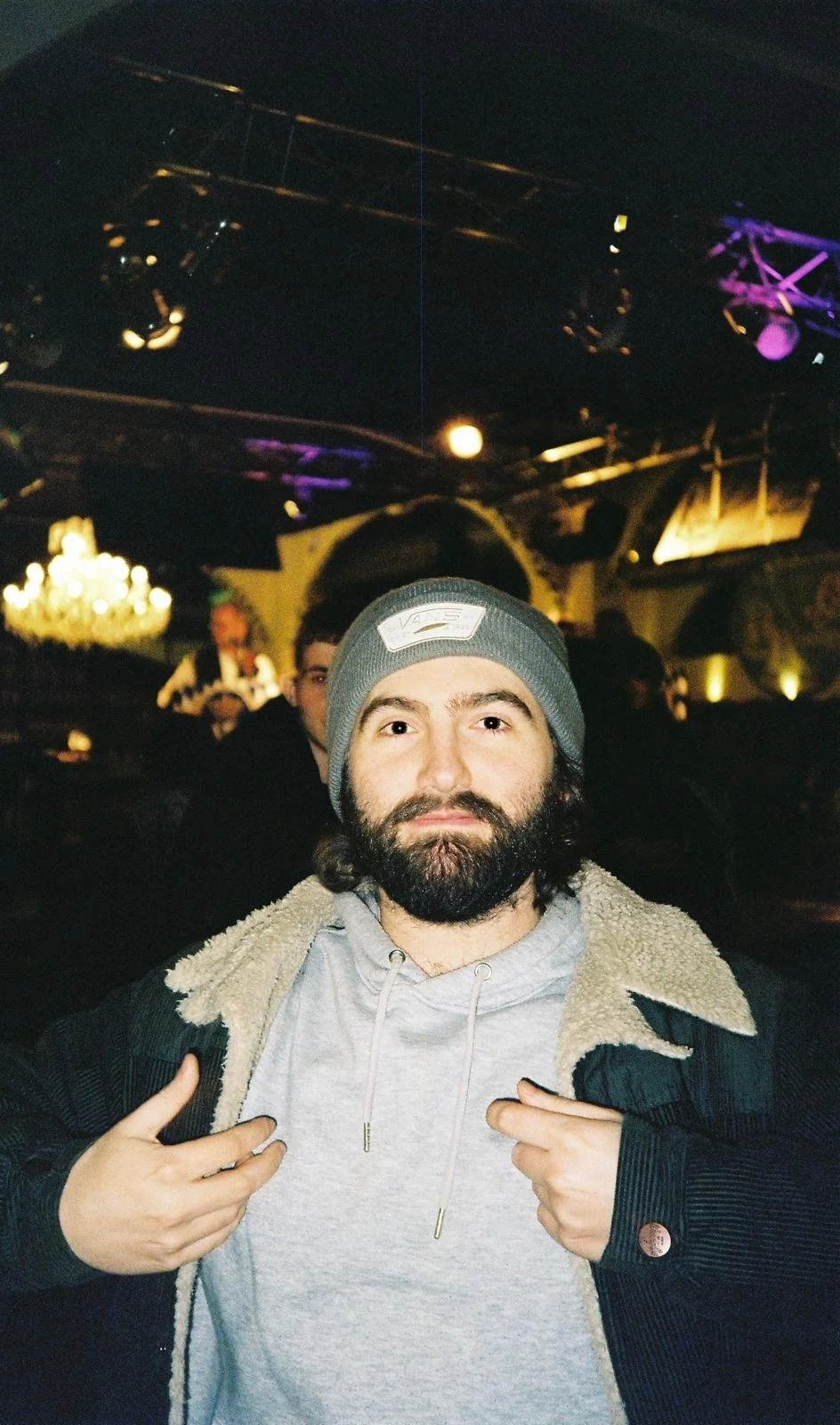 A bearded man wearing a gray beanie and gray hoodie, with a black jacket that has a shearling collar, posing indoors with a tattooed hand pointing at himself. The background shows a dimly lit room with other people, purple lighting, and a chandelier.