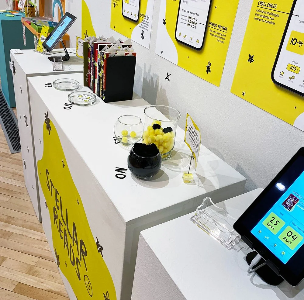 Side view of Stellar Reads exhibit with app posters, tablets, books, and jars filled with yellow and black pom-poms.