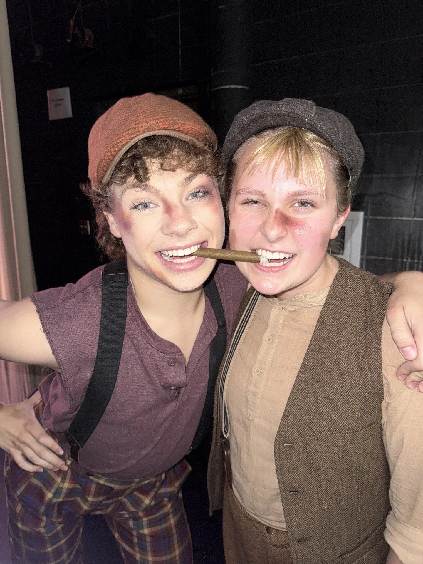 Two people in vintage costumes posing with a prop, one holding a cigar and both smiling, in a dark indoor setting.