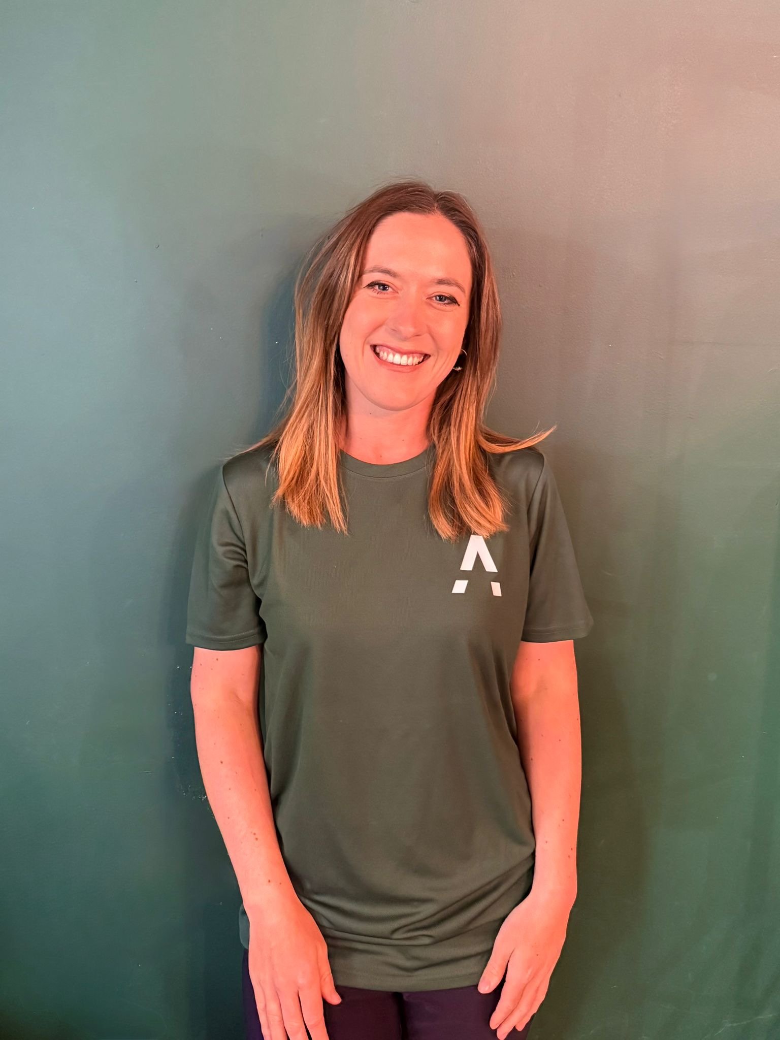 Smiling woman with shoulder-length hair wearing an olive green T-shirt standing against a plain green wall.