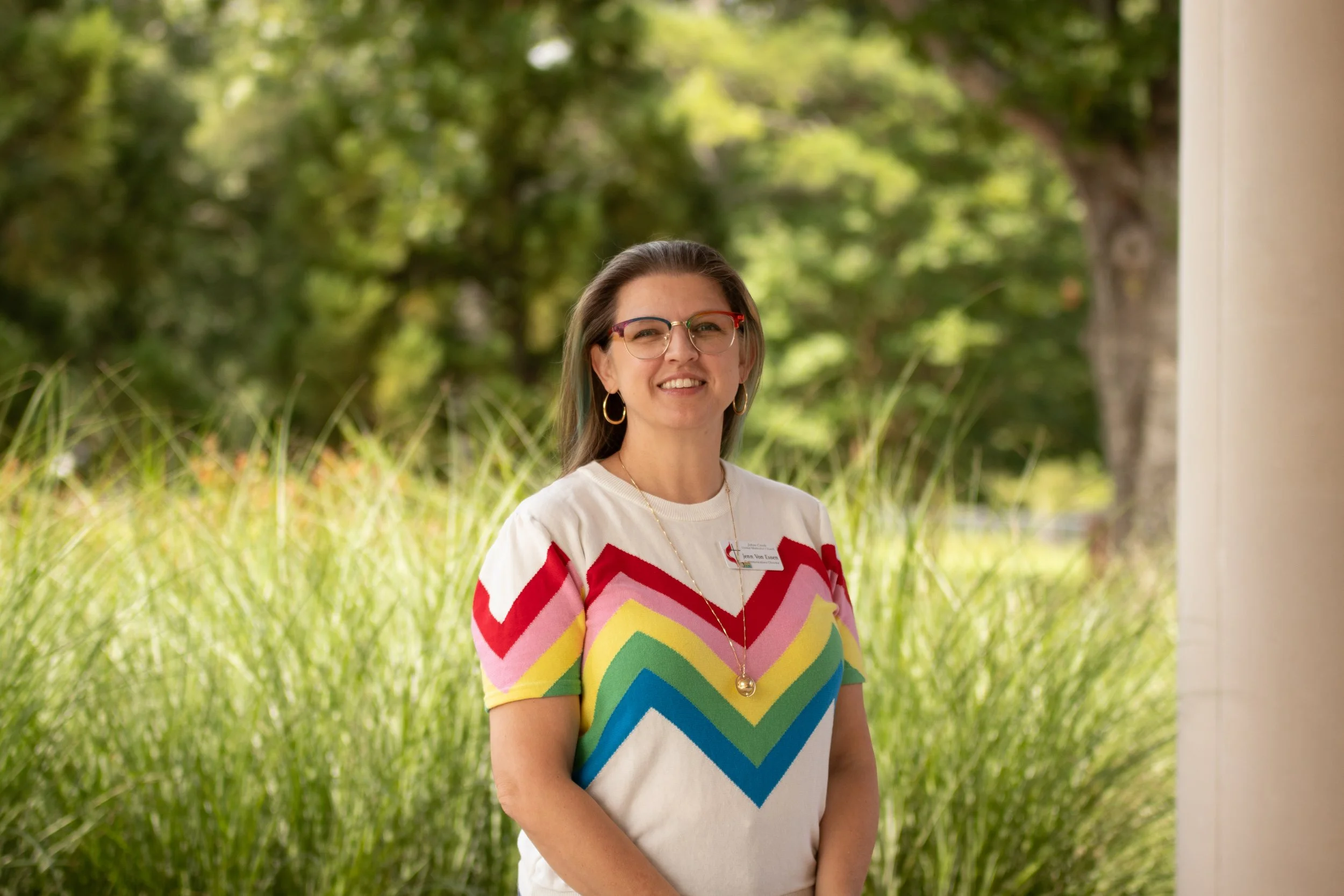 A woman with brown hair, wearing glasses and a rainbow top, standing in front tall grass.