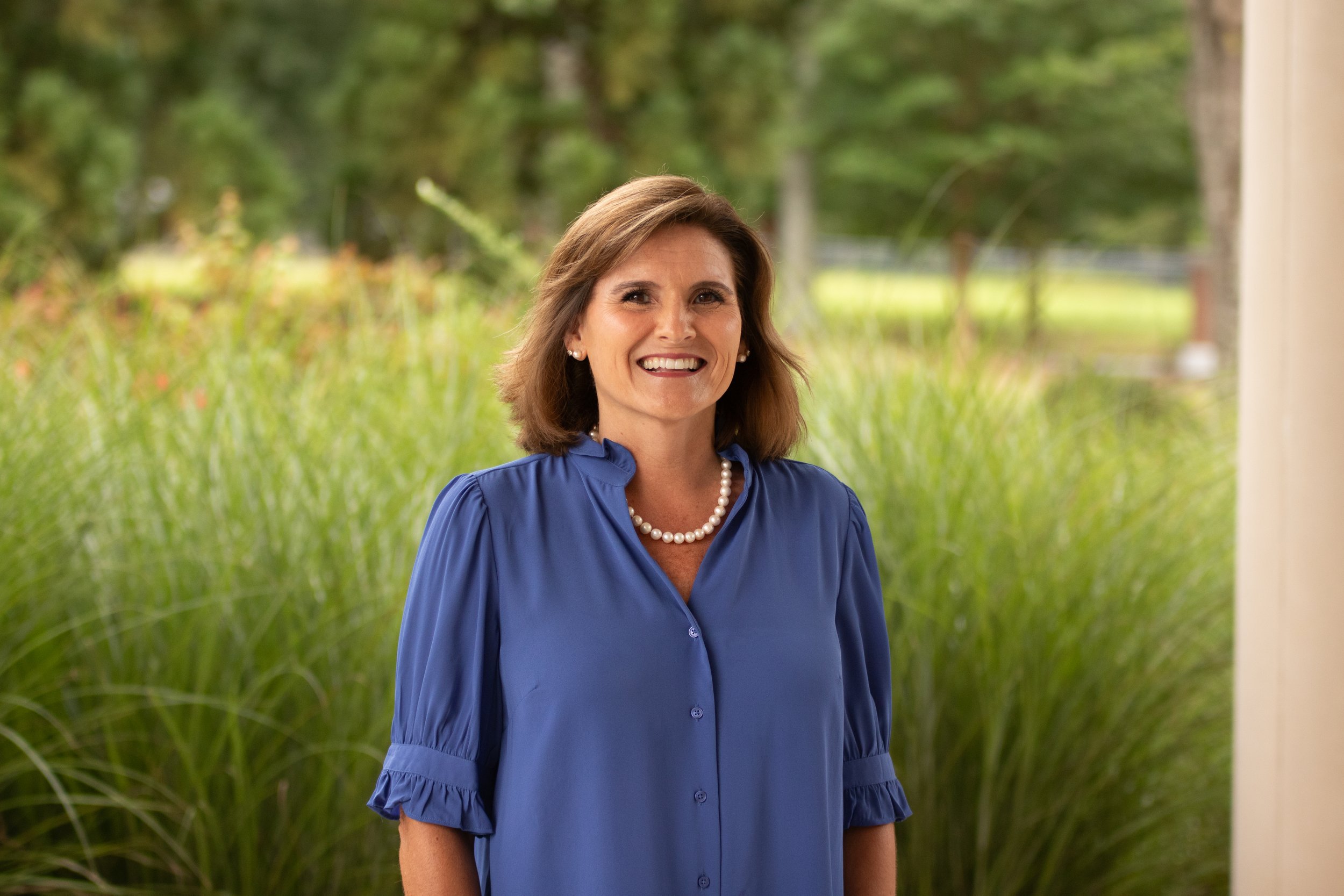 Close-up photo of a woman with dark hair, smiling, wearing a blue top, jewelry including earrings and a necklace, standing in front of tall grass.