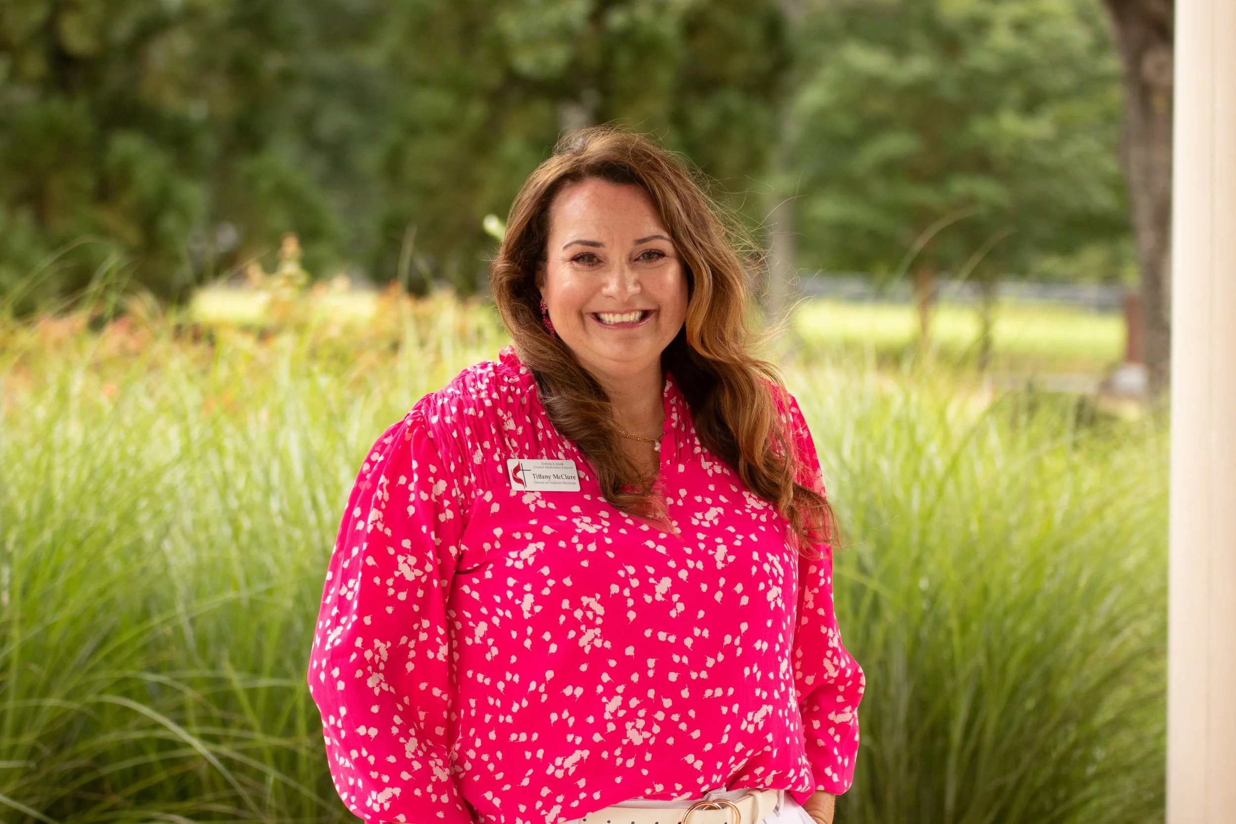 A woman with long brown hair smiling in front of a decorated Christmas tree wearing a green shirt with white text and a silver cross necklace.