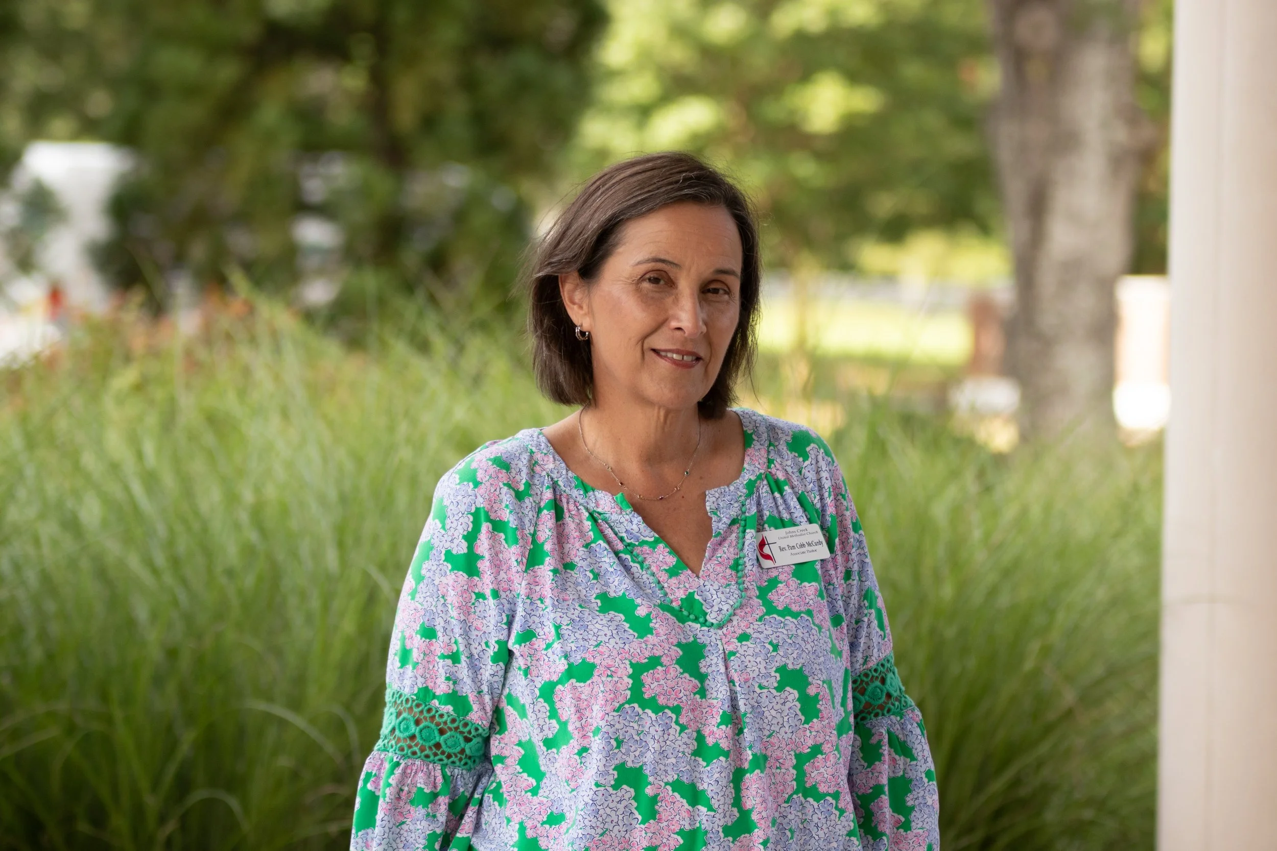 Woman in floral shirt standing in front of tall grass smiling.