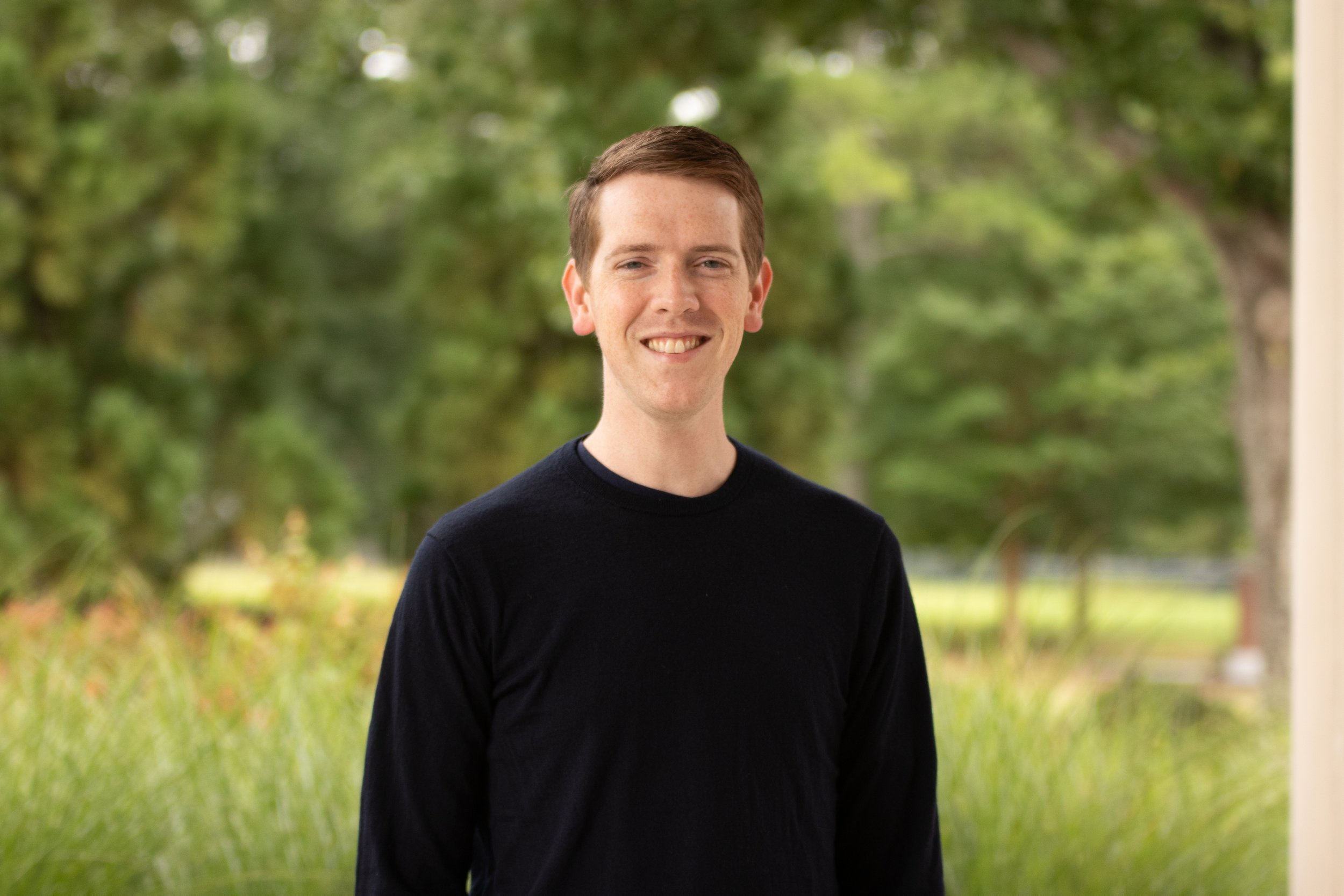 A young man with short brown hair, wearing a dark blue checked shirt, smiling and standing with arms crossed outdoors in front of green foliage.