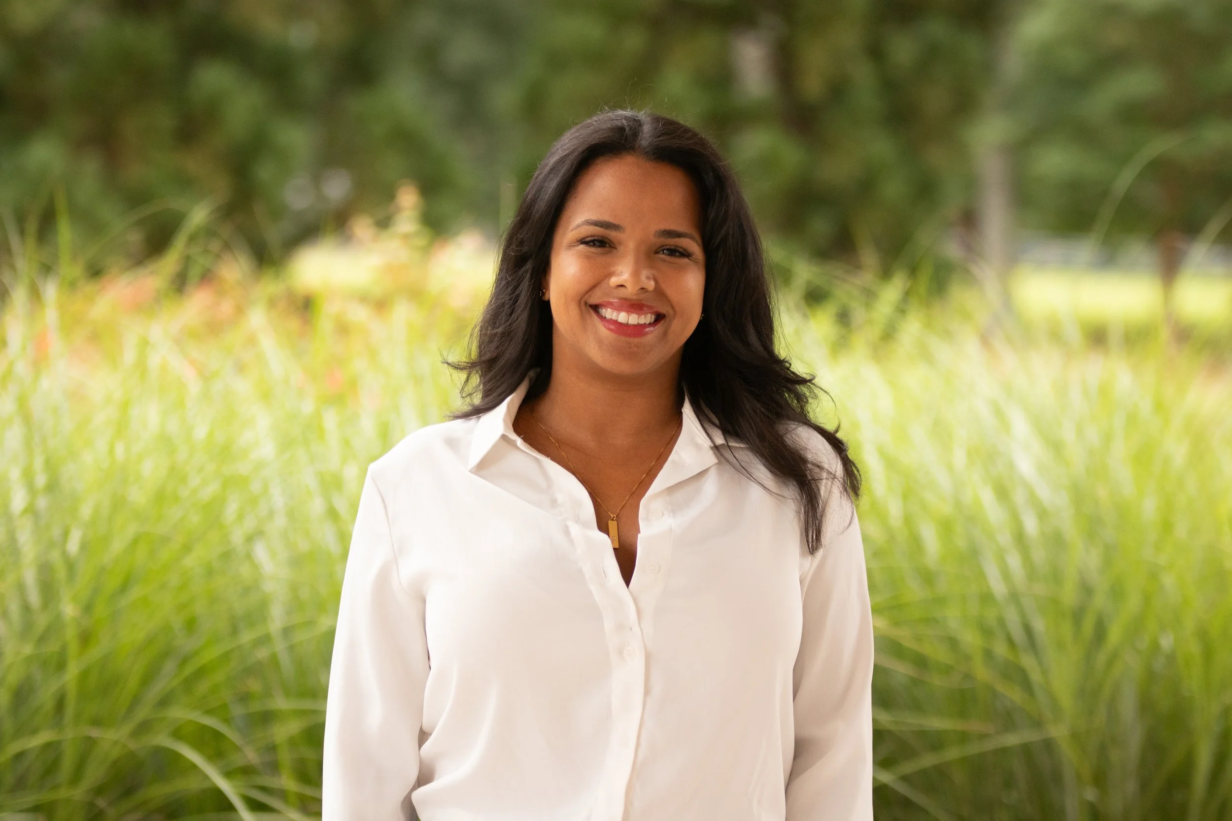 A woman with long dark hair smiling outdoors with colorful fall foliage in the background.