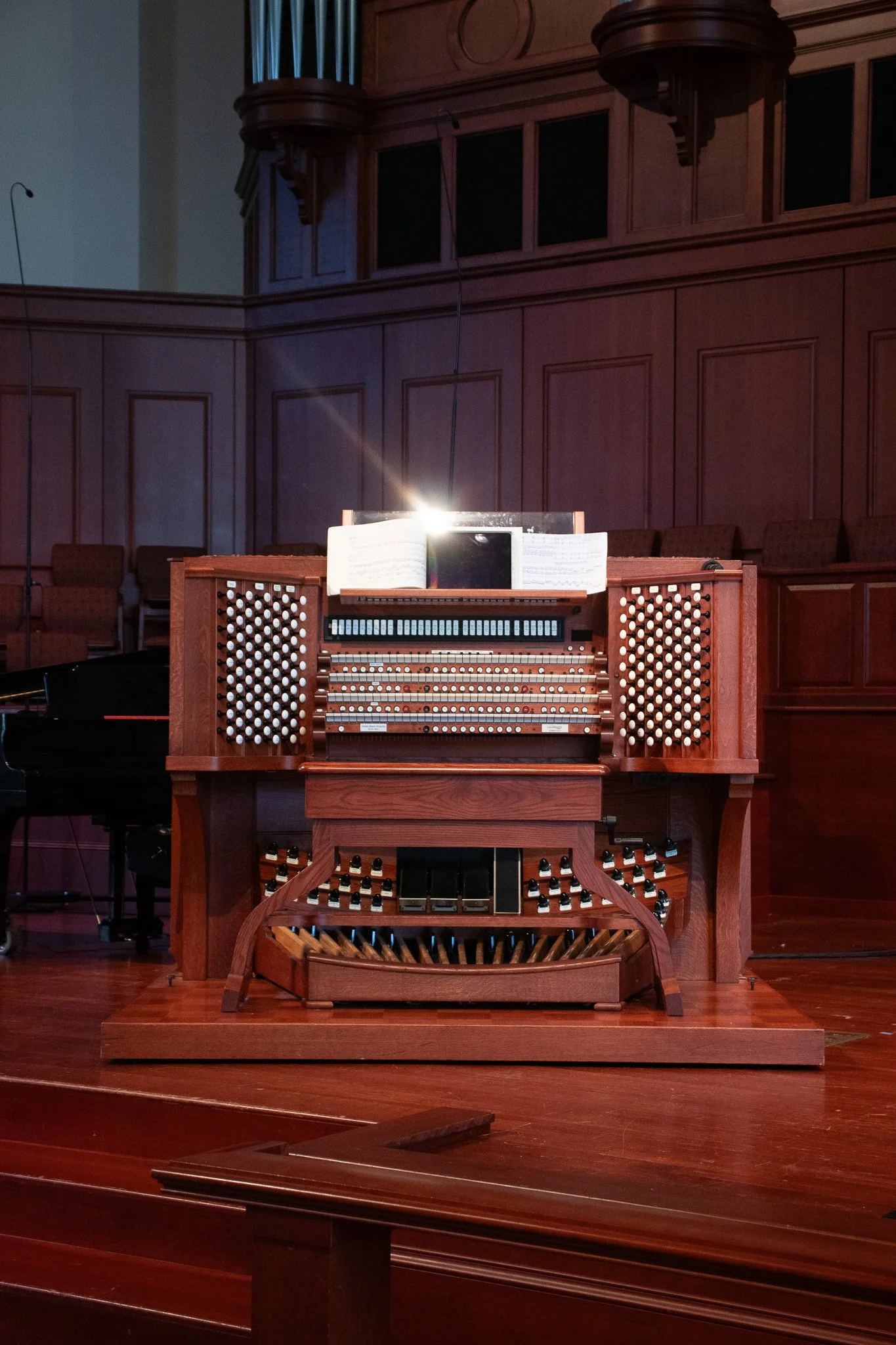 A pipe organ on a stage in a church or concert hall, with wood paneling on the wall behind it and music sheets on its top.
