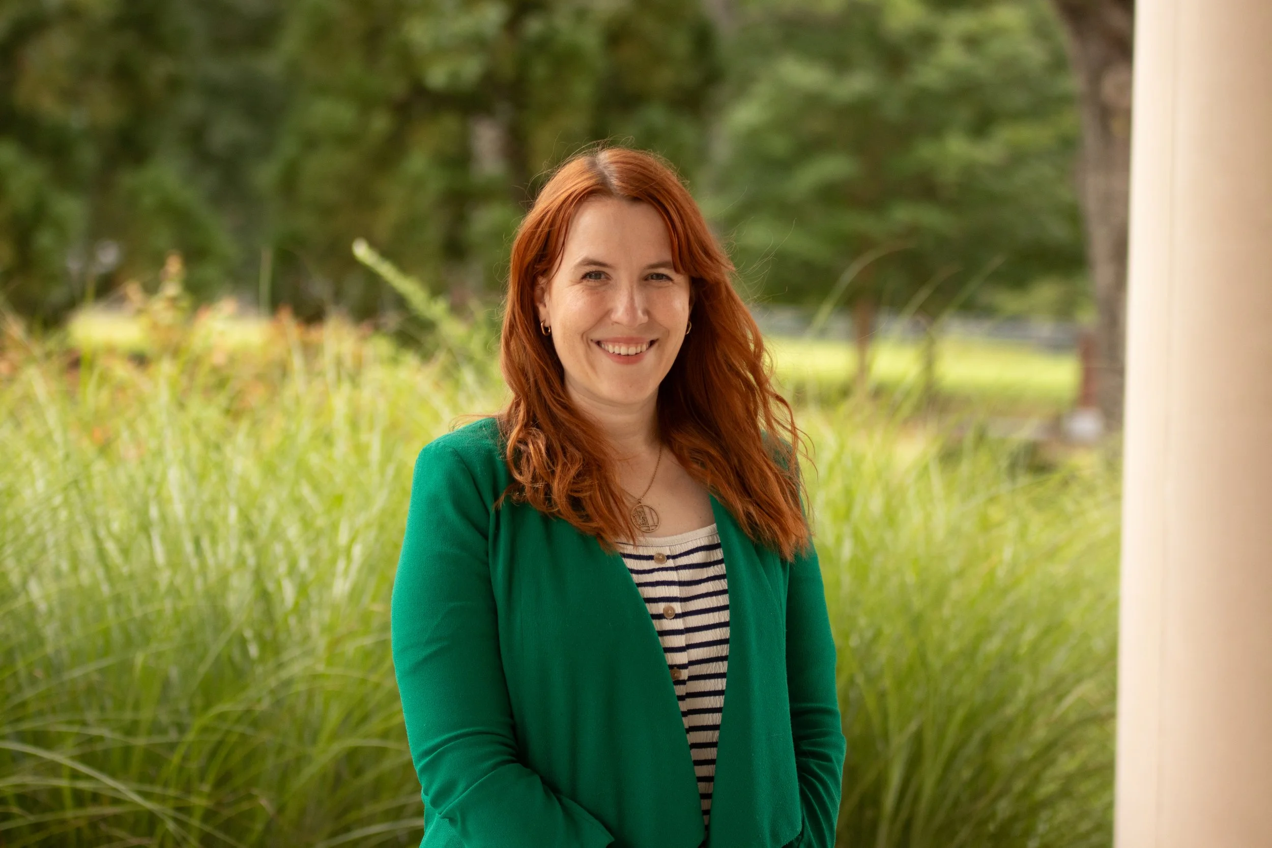 A woman with long red hair and a friendly smile, wearing a light green blazer over a white shirt, standing outdoors in front of a blurred background of stairs and greenery.
