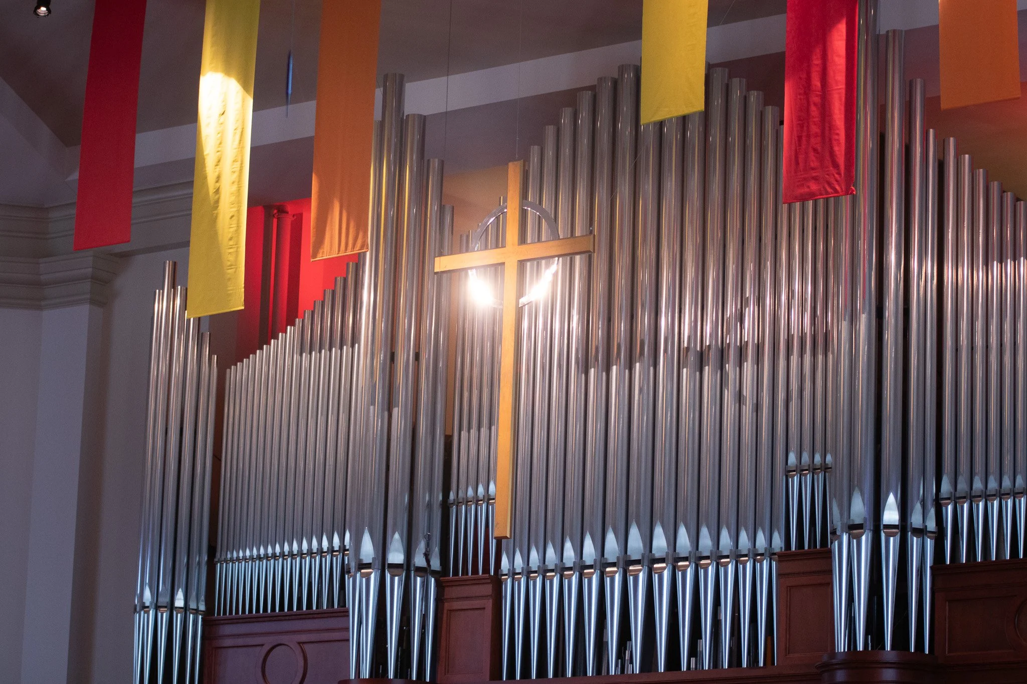 A church pipe organ with an illuminated cross hanging above it, decorated with colorful hanging banners in yellow, orange, and red.