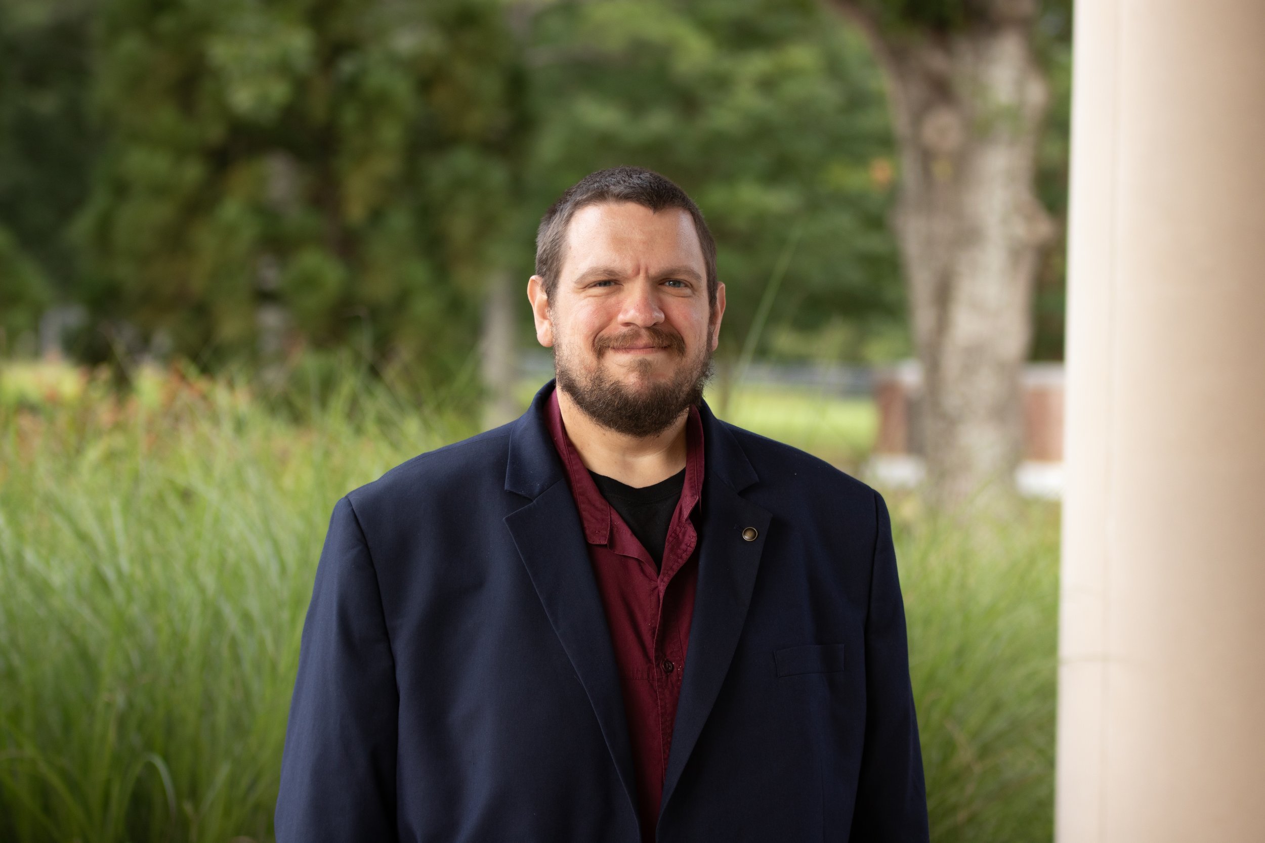 A man with dark hair and a beard, wearing a blue sport coat and a maroon shirt, standing in front of tall grass.