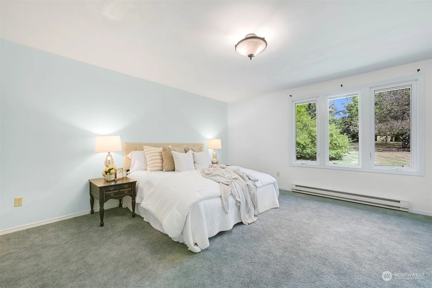 Bright bedroom with light blue accent wall, white bedding, nightstand with lamps, and large window showing greenery outside.