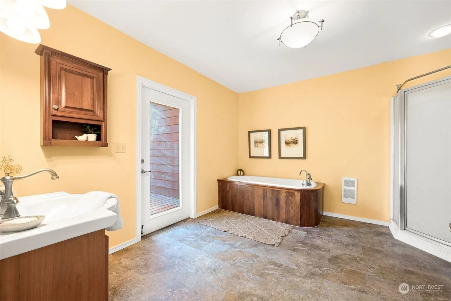 Bathroom with yellow walls, a bathtub with wooden paneling, a walk-in shower, a small sink with a wooden cabinet, a glass door leading outside, and framed pictures on the wall.