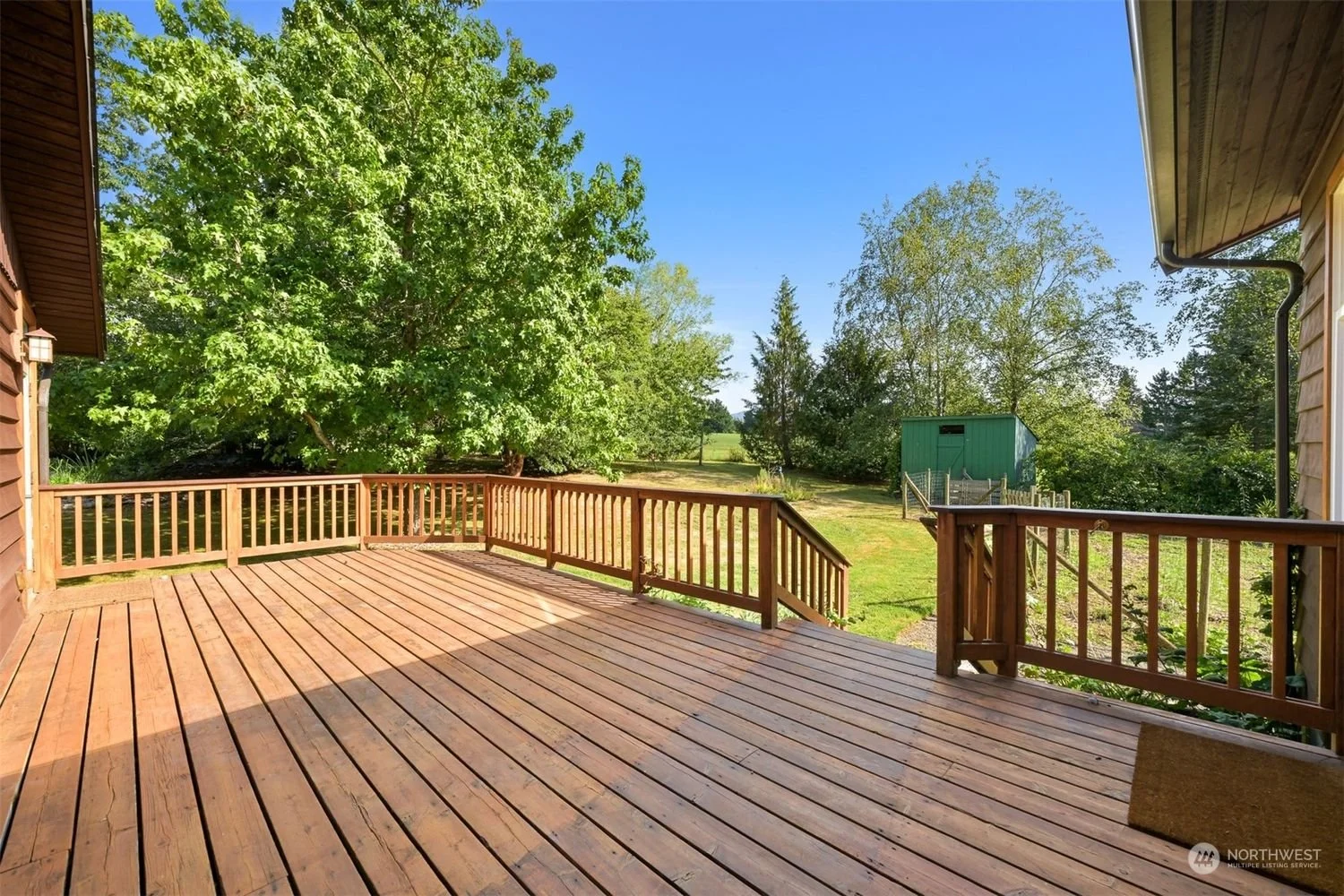 View of a wooden deck with a railing, overlooking a grassy yard with trees and a small green shed in the background on a sunny day.