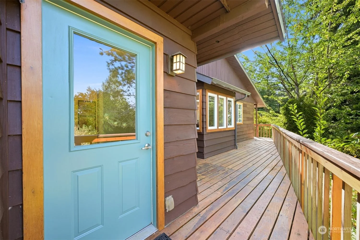 Exterior view of a house with a wooden deck, brown siding, a blue door with a window, outdoor wall light, and surrounding green trees.
