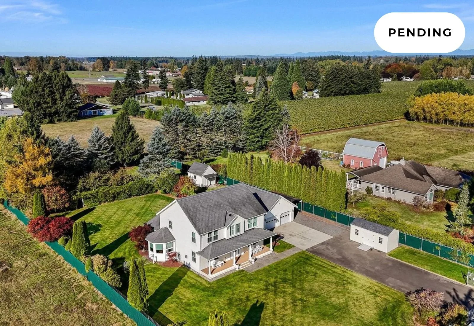 Aerial view of a suburban neighborhood with a white two-story house with a porch, a gray roof, and a green backyard, surrounded by trees and a green fence, with other houses, trees, and fields in the background under a blue sky labeled 'ACTIVE'.