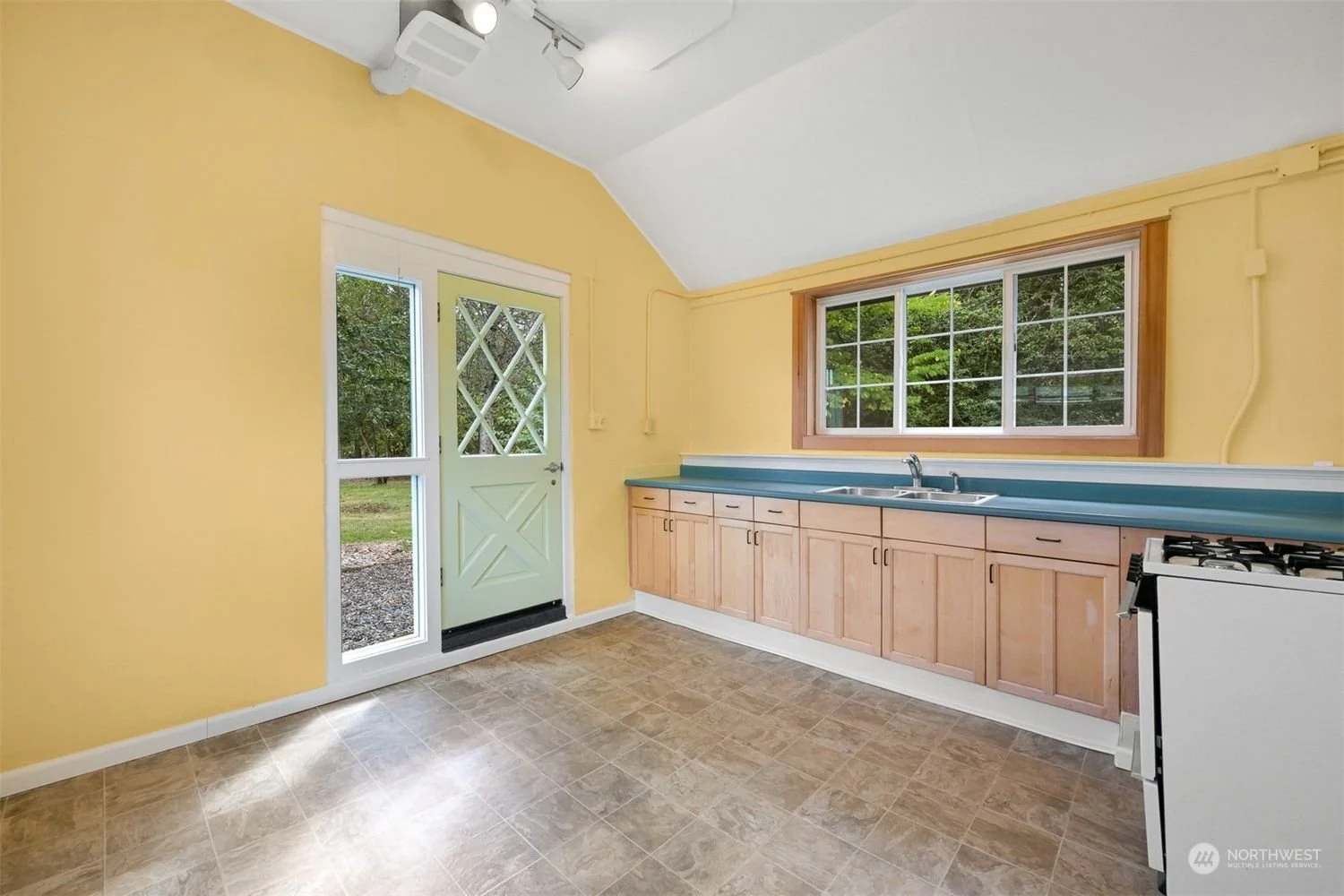 Empty kitchen with yellow walls, wooden cabinets, blue countertop, large window, tiled floor, and light green door leading outside.