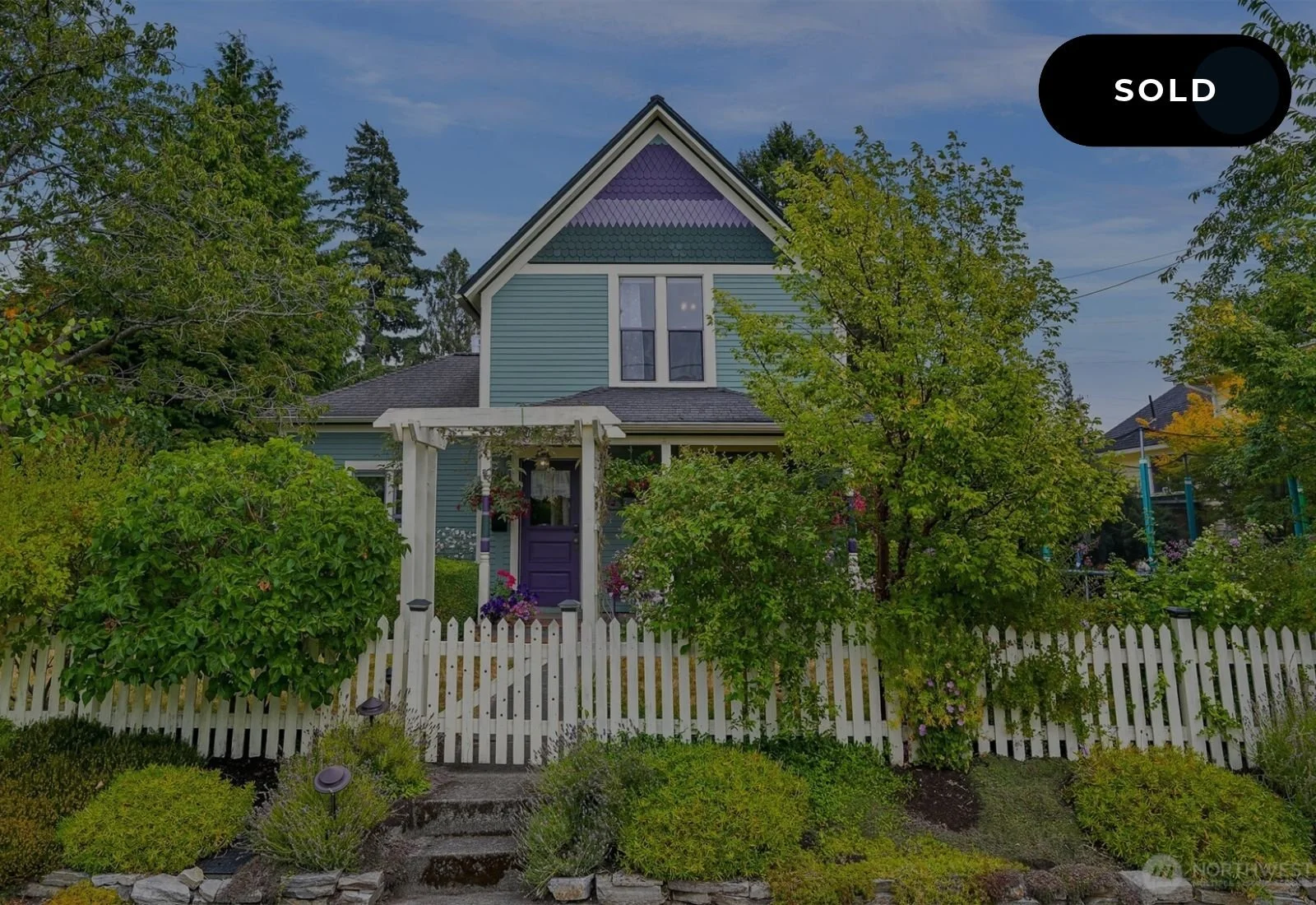 A charming two-story house with blue siding, purple accents, and a white picket fence, surrounded by lush green trees and plants.