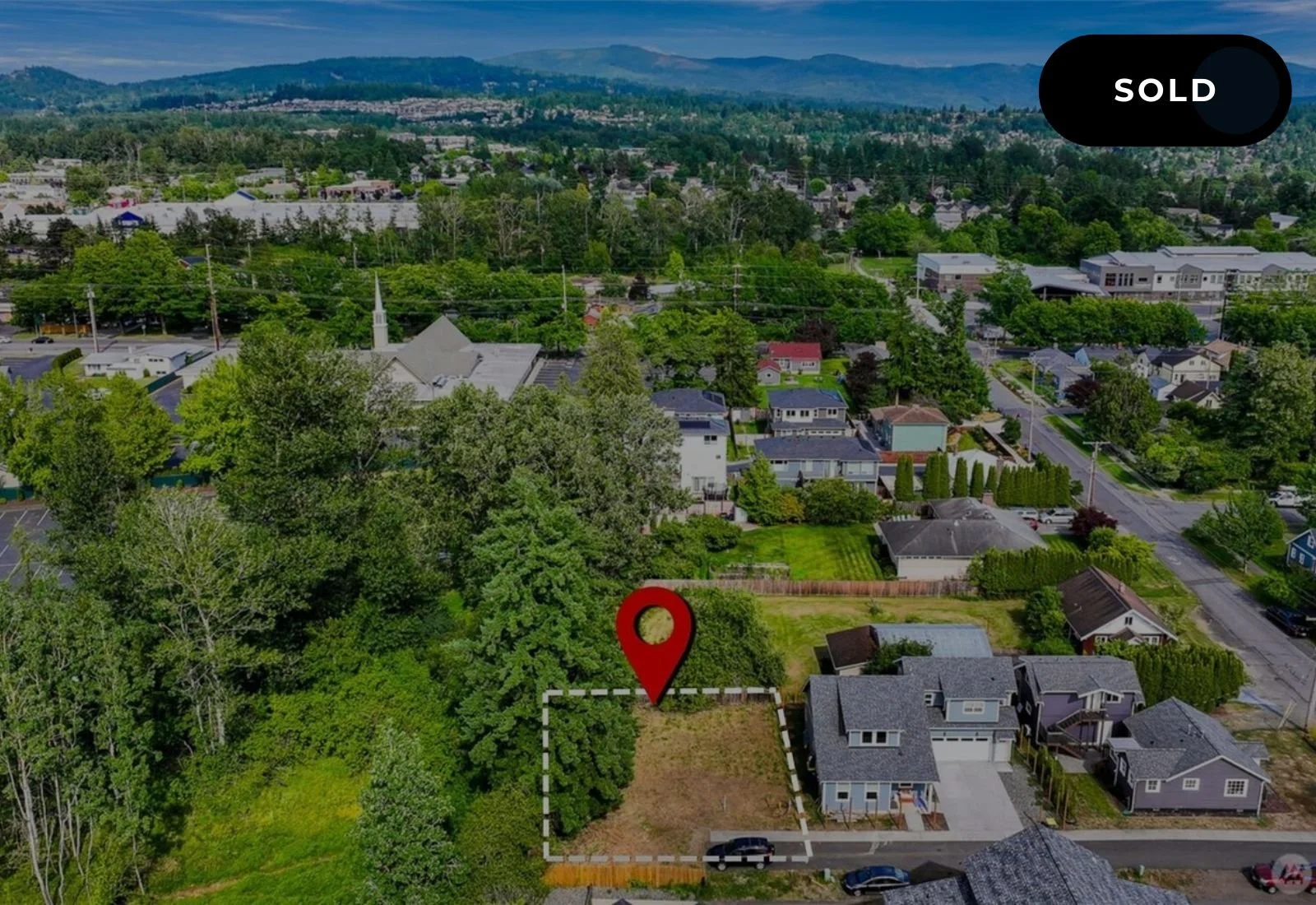 An aerial view of a residential neighborhood with a large green backyard, marked with a red location pin in the yard, and a nearby house. The image shows a suburban area with trees, houses, roads, and distant hills under a partly cloudy sky.