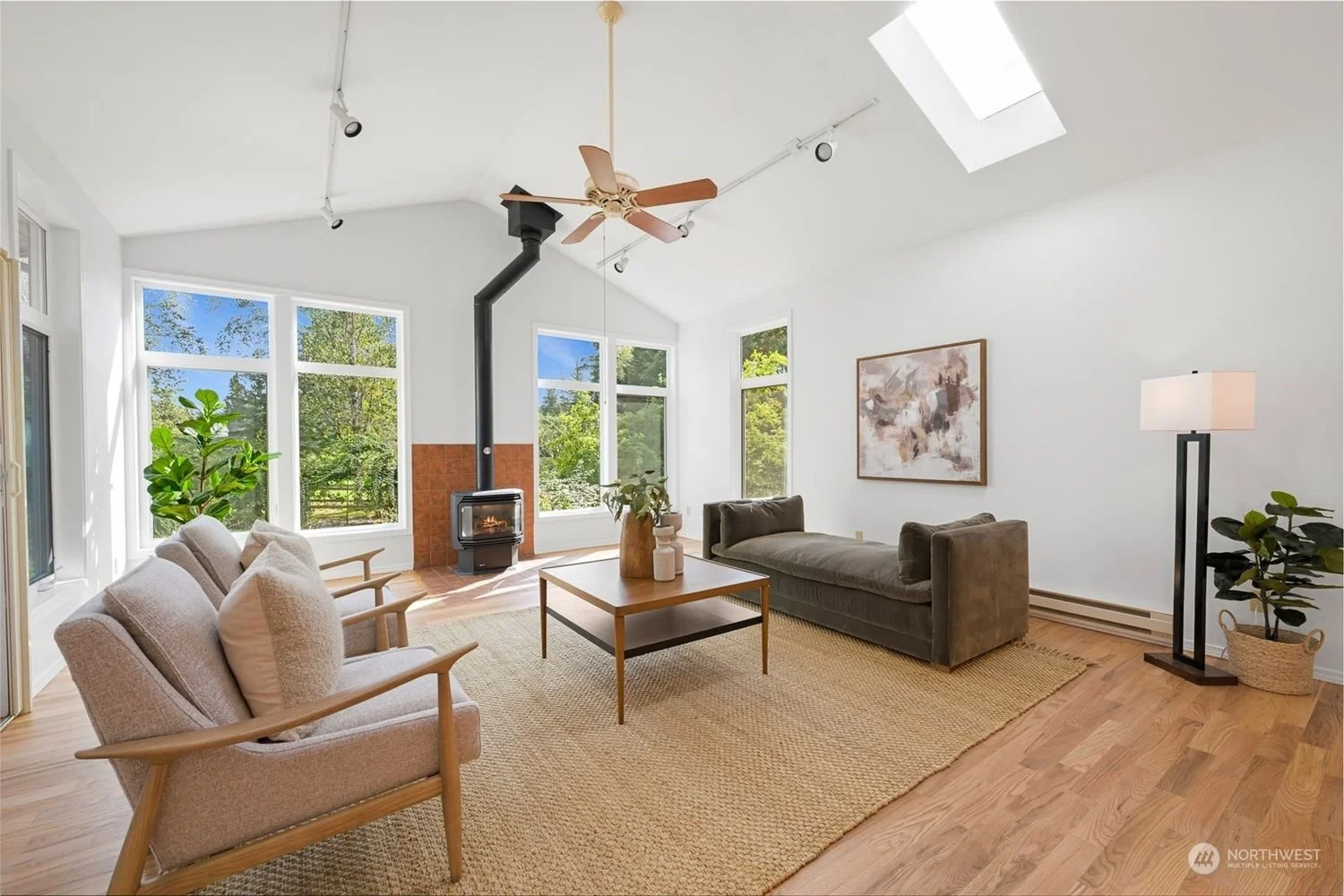 Living room with large windows, a wood-burning stove, beige and green couches, a wooden coffee table, green plants, a floor lamp, and a skylight, with white walls and hardwood floors.