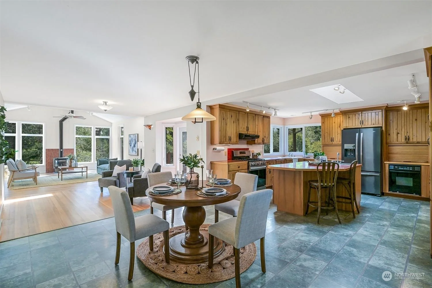 Open-concept kitchen and living room with wooden cabinets, gray refrigerator, dining table with six chairs, and a living area with couches, armchairs, a wood stove, and large windows showing greenery outside.