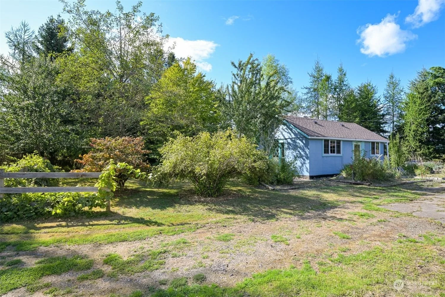 A small blue house surrounded by trees and bushes, with a wooden fence on the left and a partly with gravel and grass in the foreground, under a blue sky with some clouds.