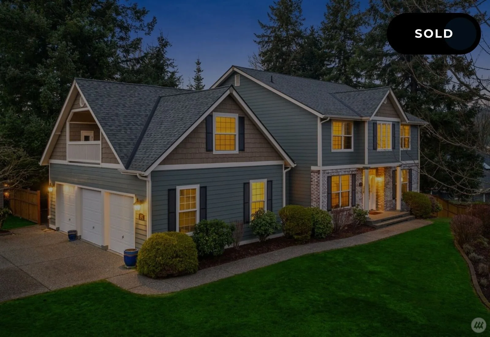 Two-story suburban house with gray siding, stone accents, and a dark roof, lit interior windows, front steps, landscaped yard with bushes, a curved walkway, and a green lawn, with a 