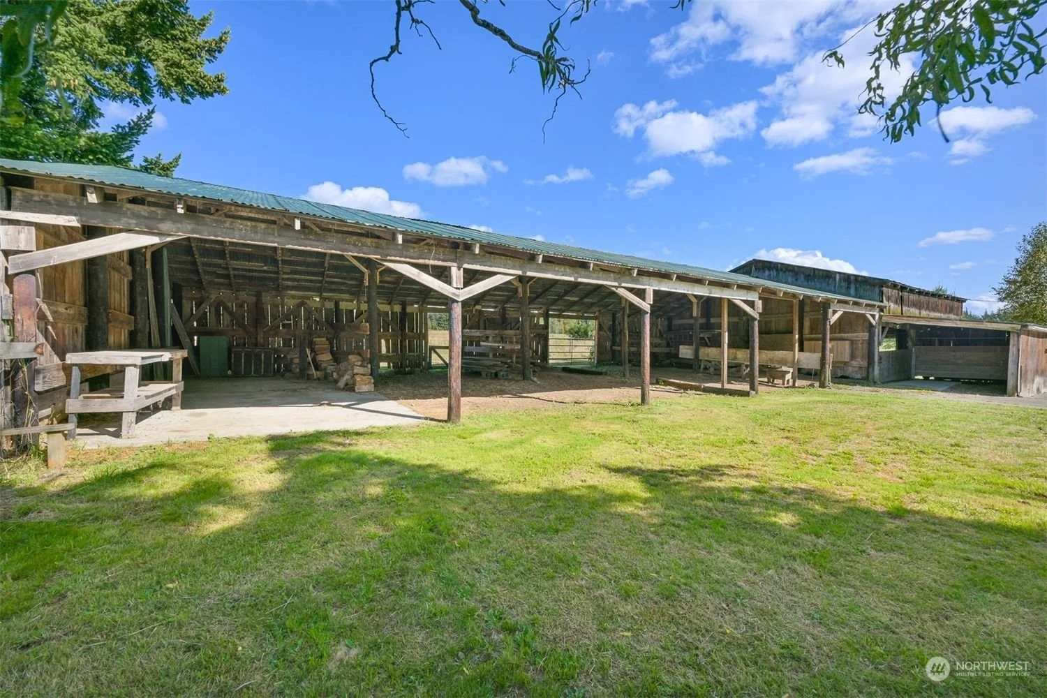 A rustic wooden barn with a corrugated metal roof, open sides, and a grassy foreground under a partly cloudy blue sky.
