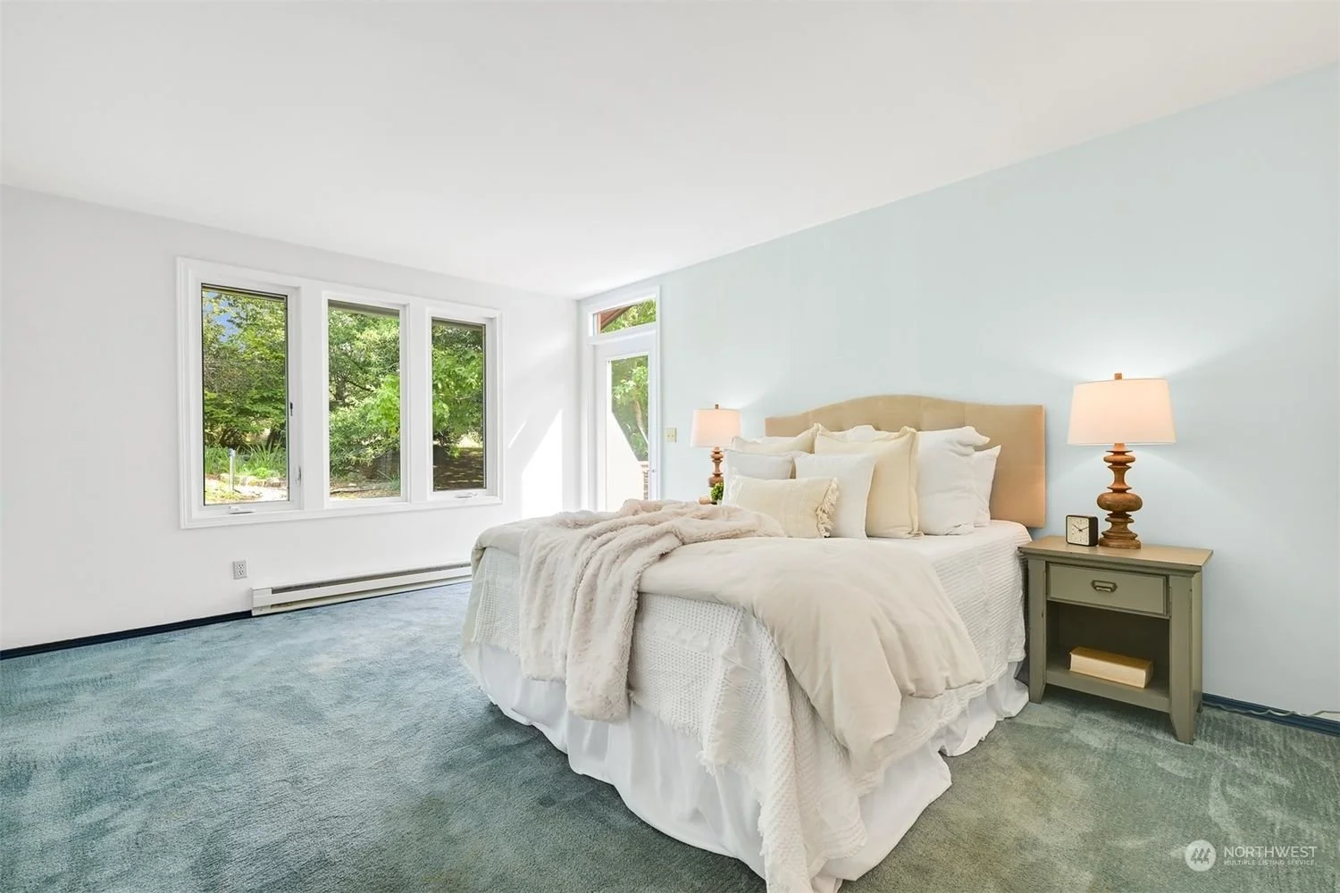A bright bedroom with a bed covered in white and beige bedding, two beige nightstands with lamps, and three large windows showing green trees outside.