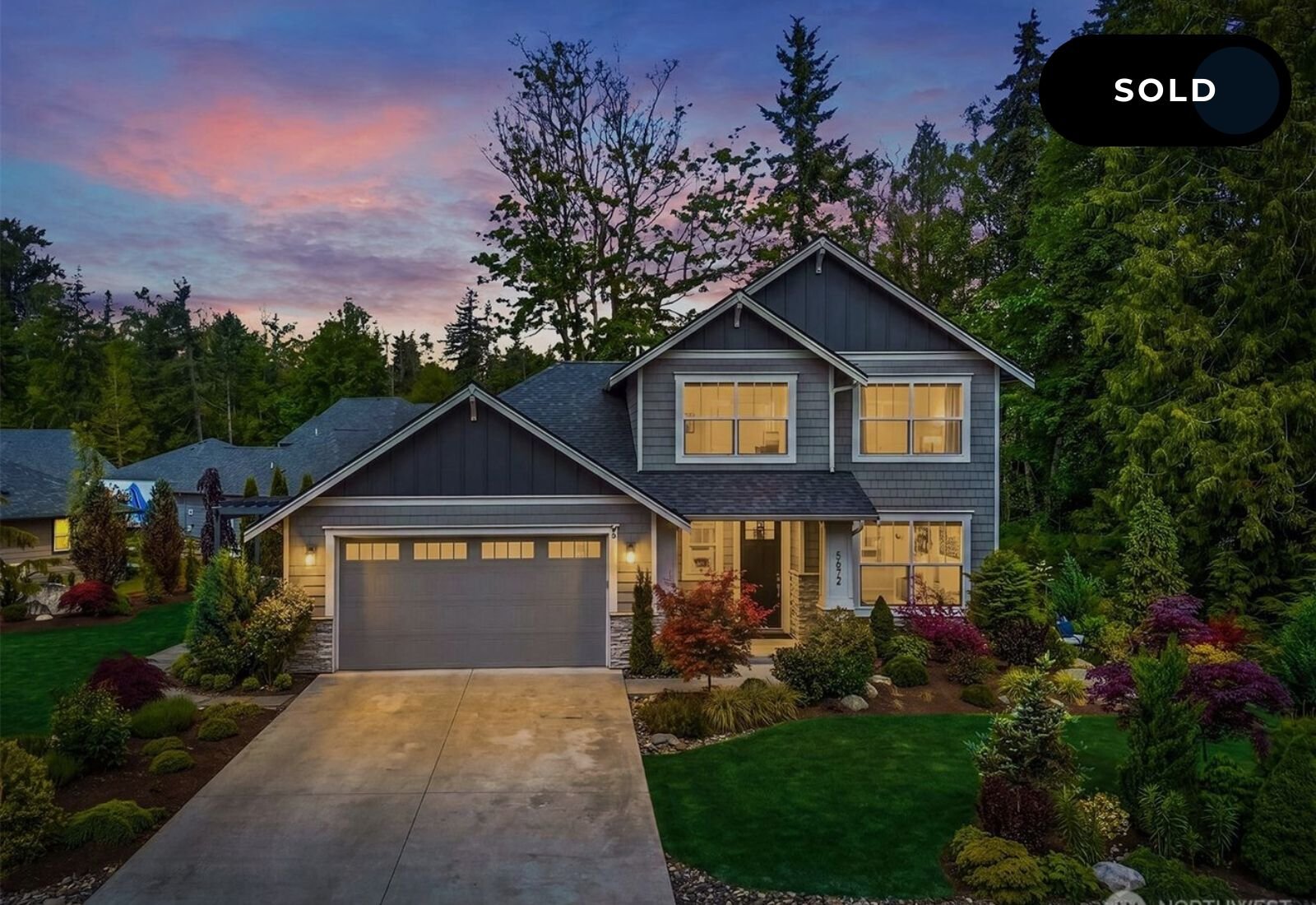 A two-story house with gray siding, illuminated windows, and a two-car garage, set in a landscaped yard with lush trees and colorful shrubs during sunset.