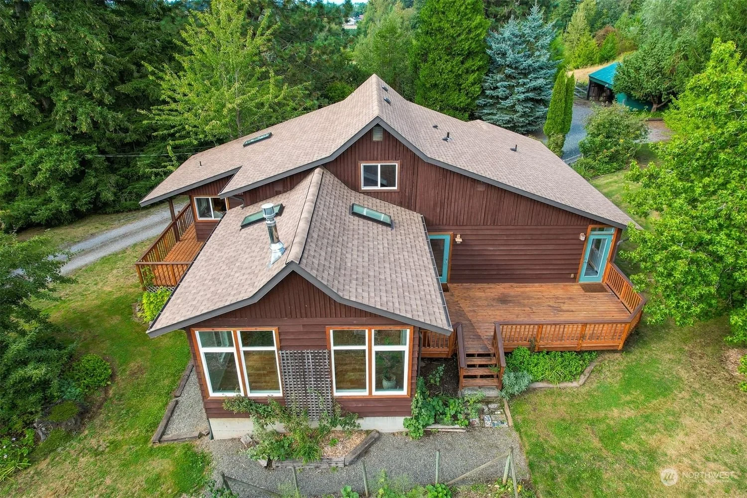 Aerial view of a two-story wooden house with multiple roof sections and skylights, surrounded by trees and greenery, featuring multiple decks with stairs and a backyard garden.