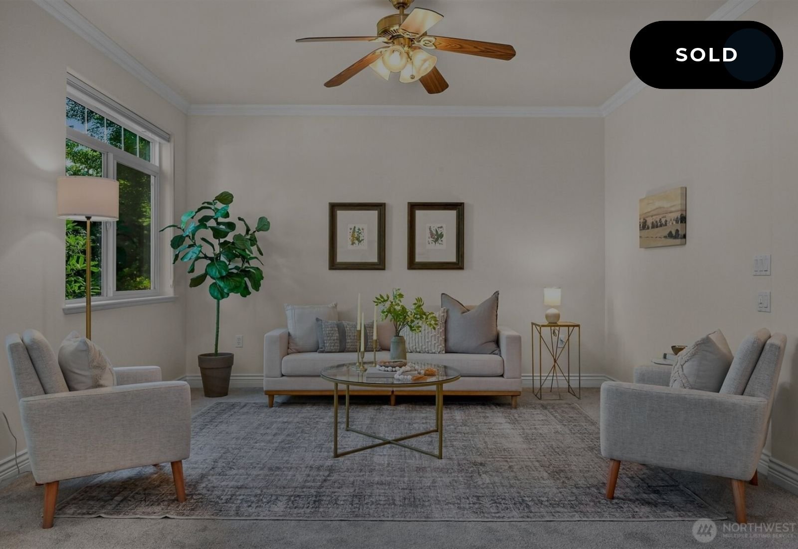 Living room with beige walls, a ceiling fan with wooden blades, a window with natural light, a large potted plant, a white sofa with decorative pillows, glass coffee table with decor, side tables with lamps, framed art on the wall, and a cozy rug.